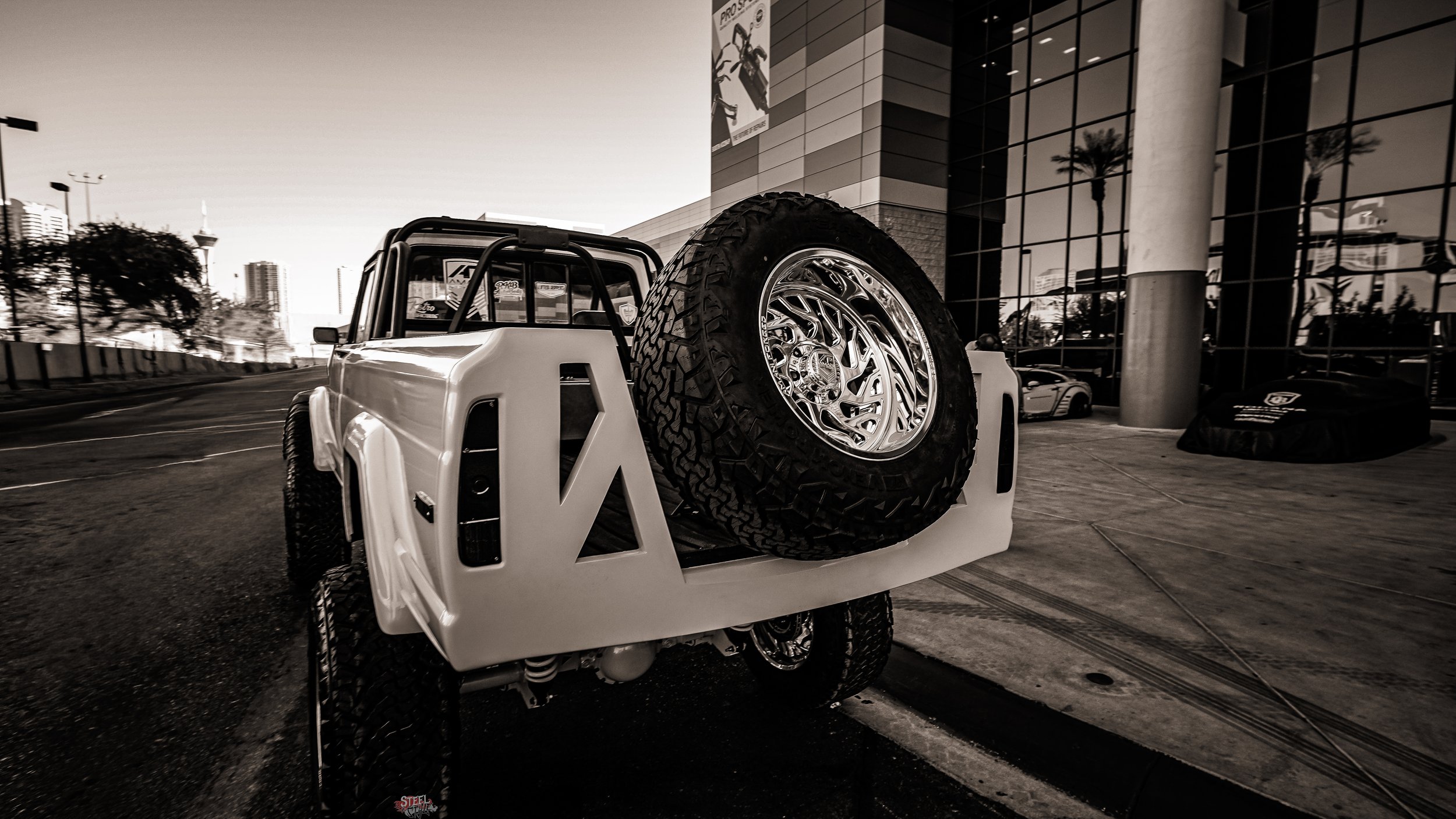A white off-road vehicle with a large tire mounted on the back, parked outside a modern building with glass windows.