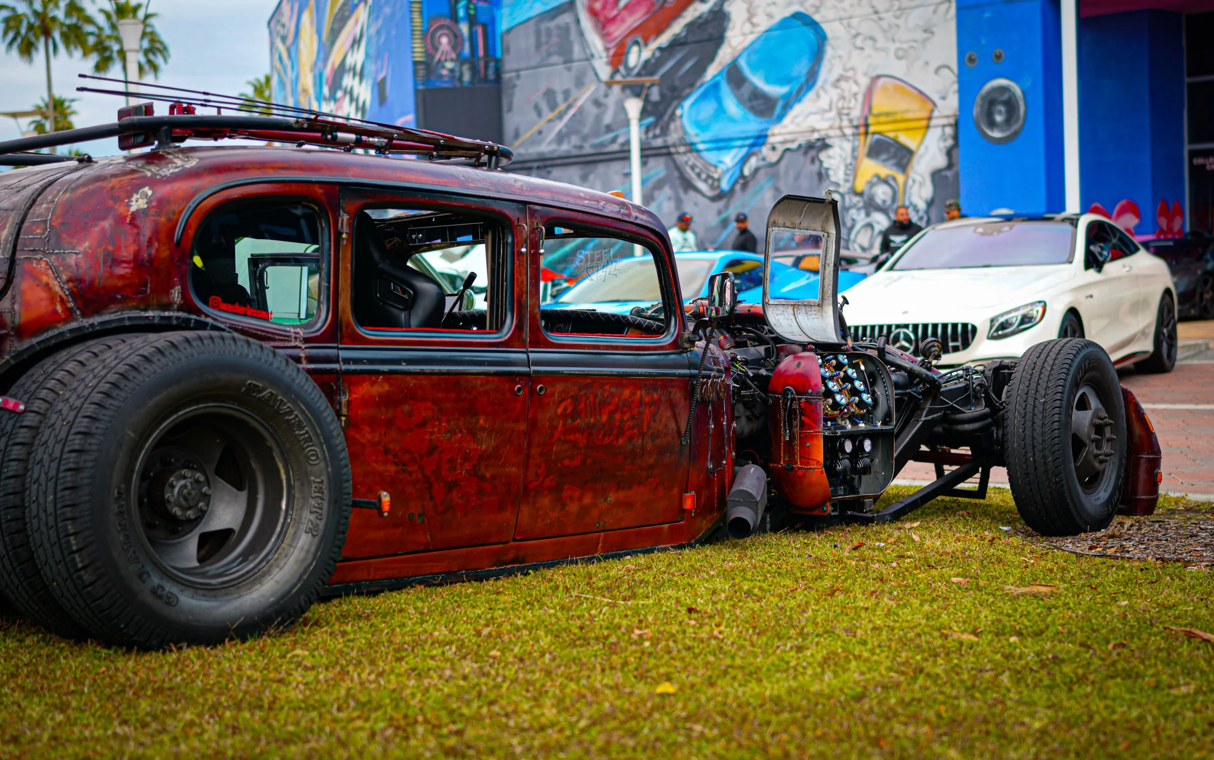 A vintage car with a rusted exterior and exposed engine parts parked on grass, with modern cars and colorful street art in the background.