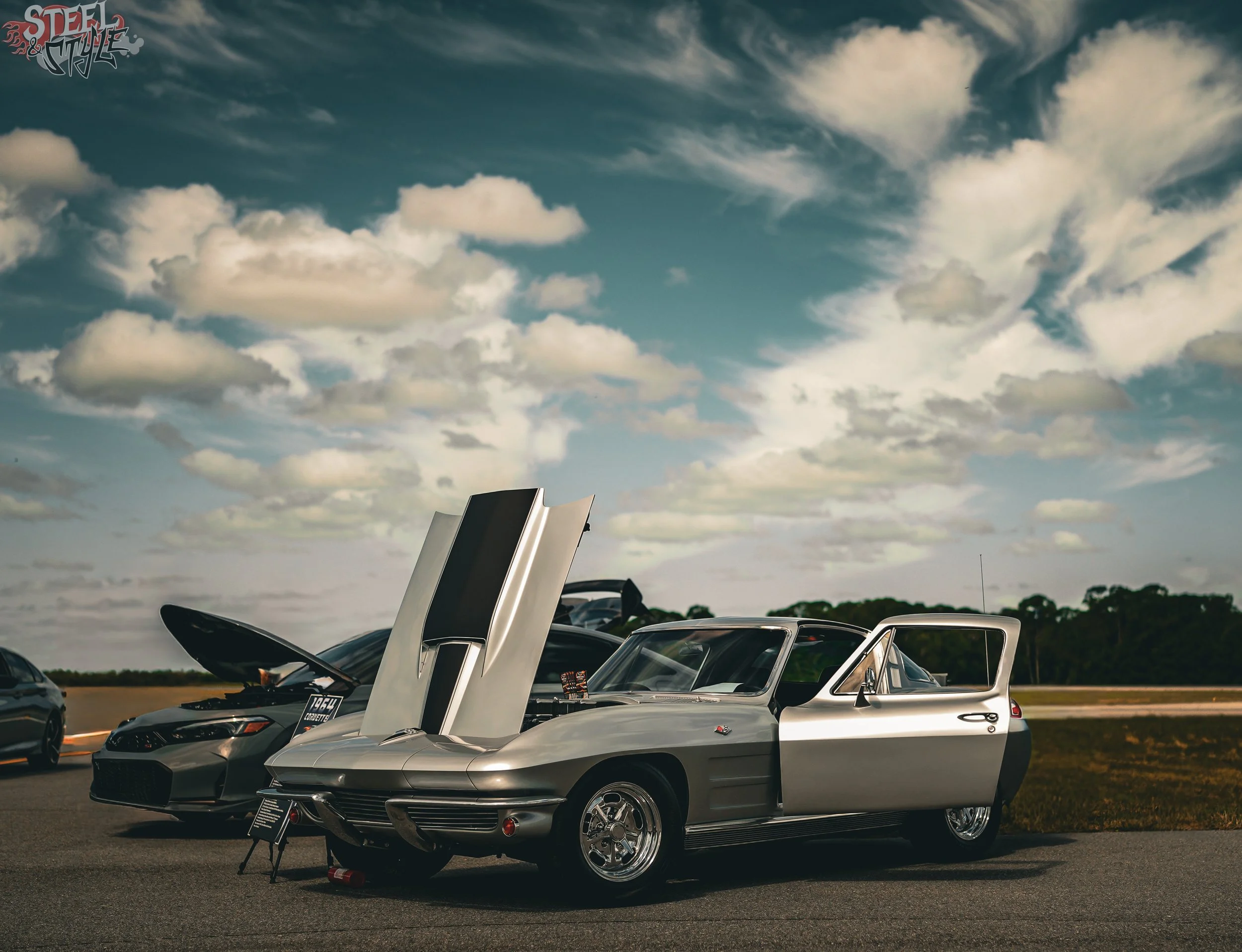 A row of classic cars on display at an outdoor car show, with their hoods open and a partly cloudy sky above