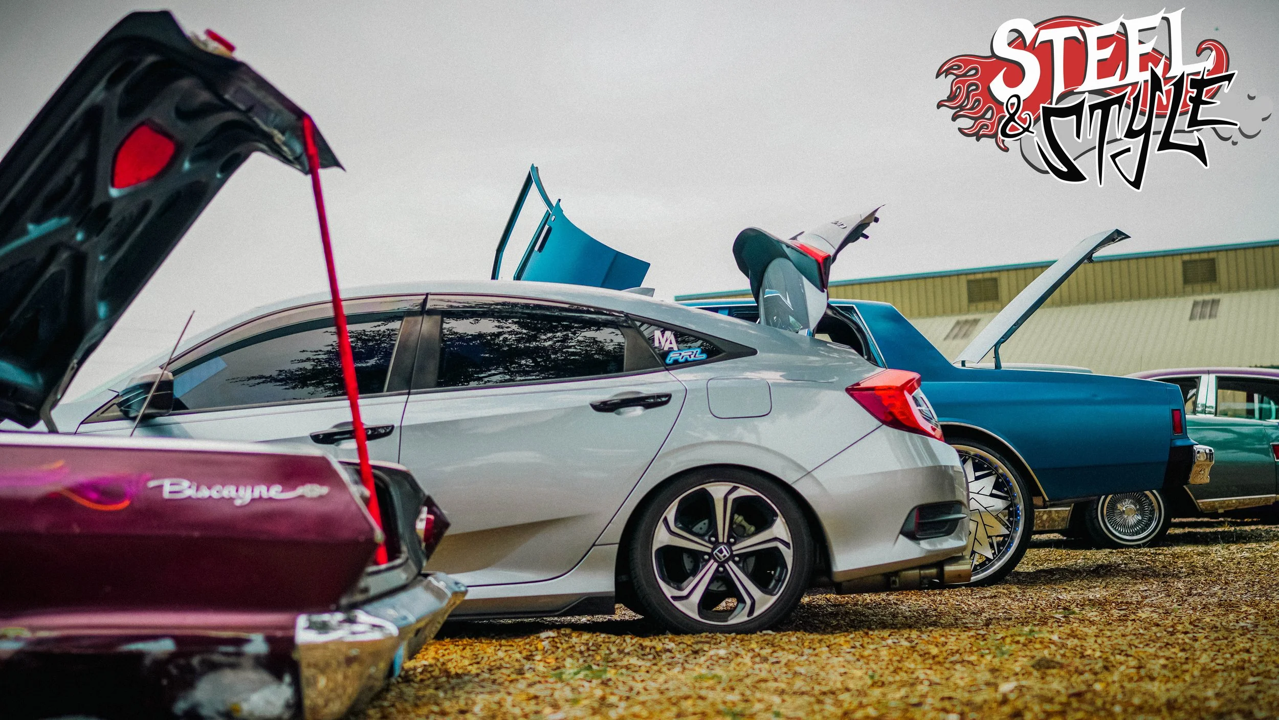 A lineup of cars parked outdoors with their hoods and trunks open. The image features a white modern compact car, a blue vintage car, and part of other cars, with the logo 'Steel & Style' in the upper right corner.