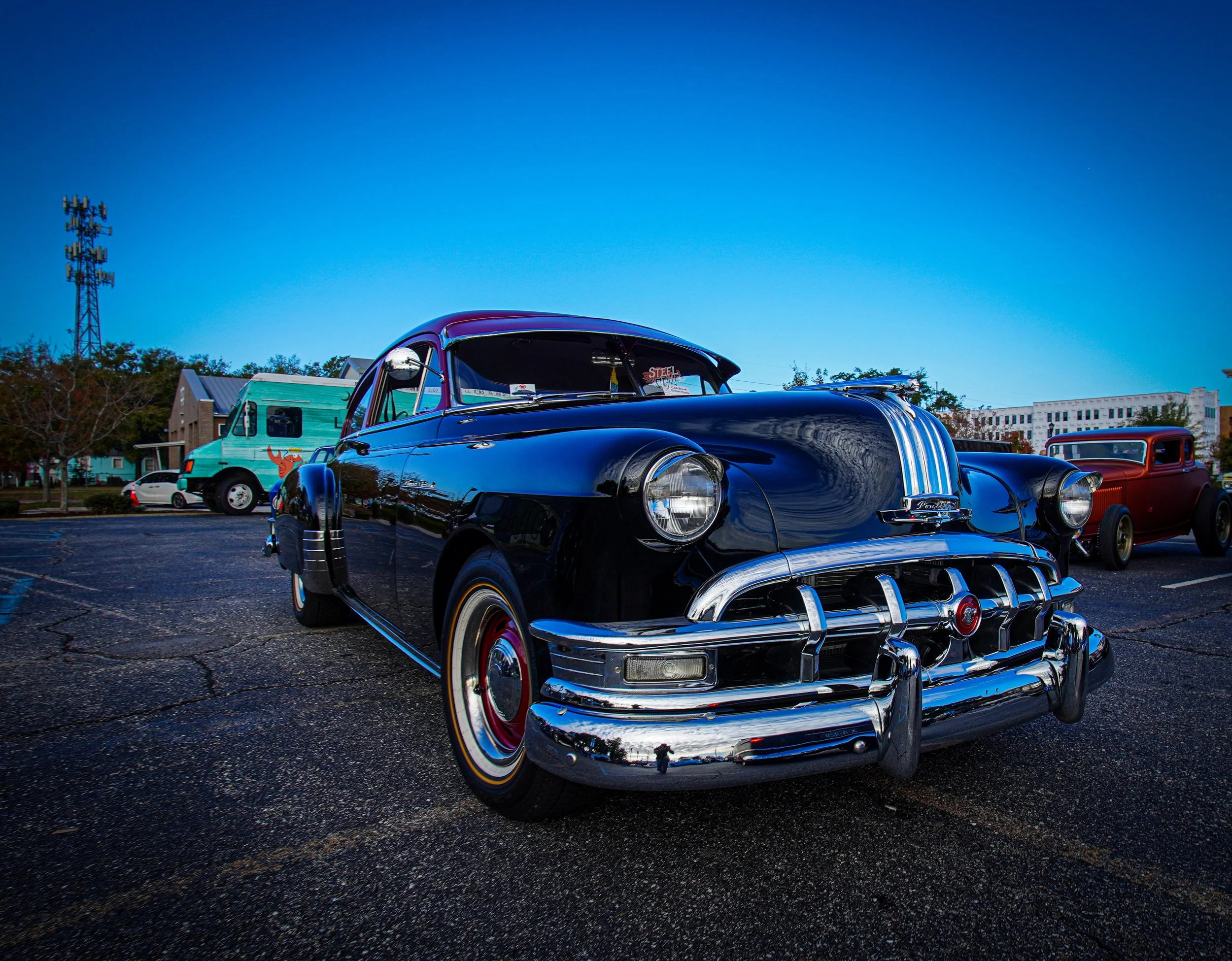 Black vintage Cadillac car parked in an outdoor lot with other classic vehicles, against a backdrop of trees, buildings, and a clear blue sky.
