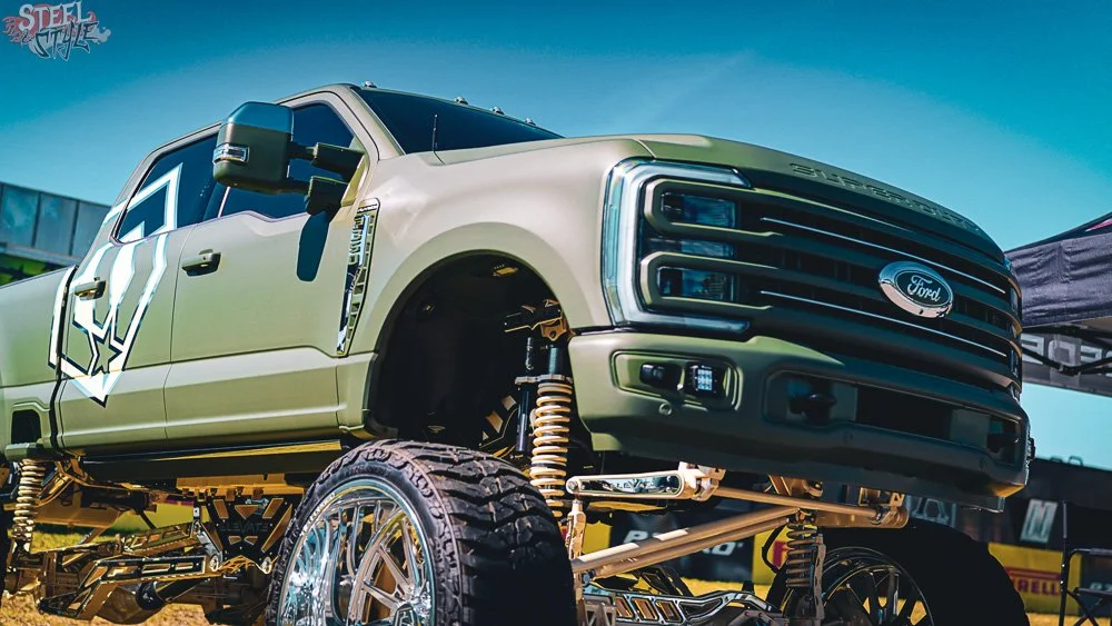 Modified Ford Super Duty truck with oversized tires, lifted suspension, and custom bodywork, parked outdoors at a car event.