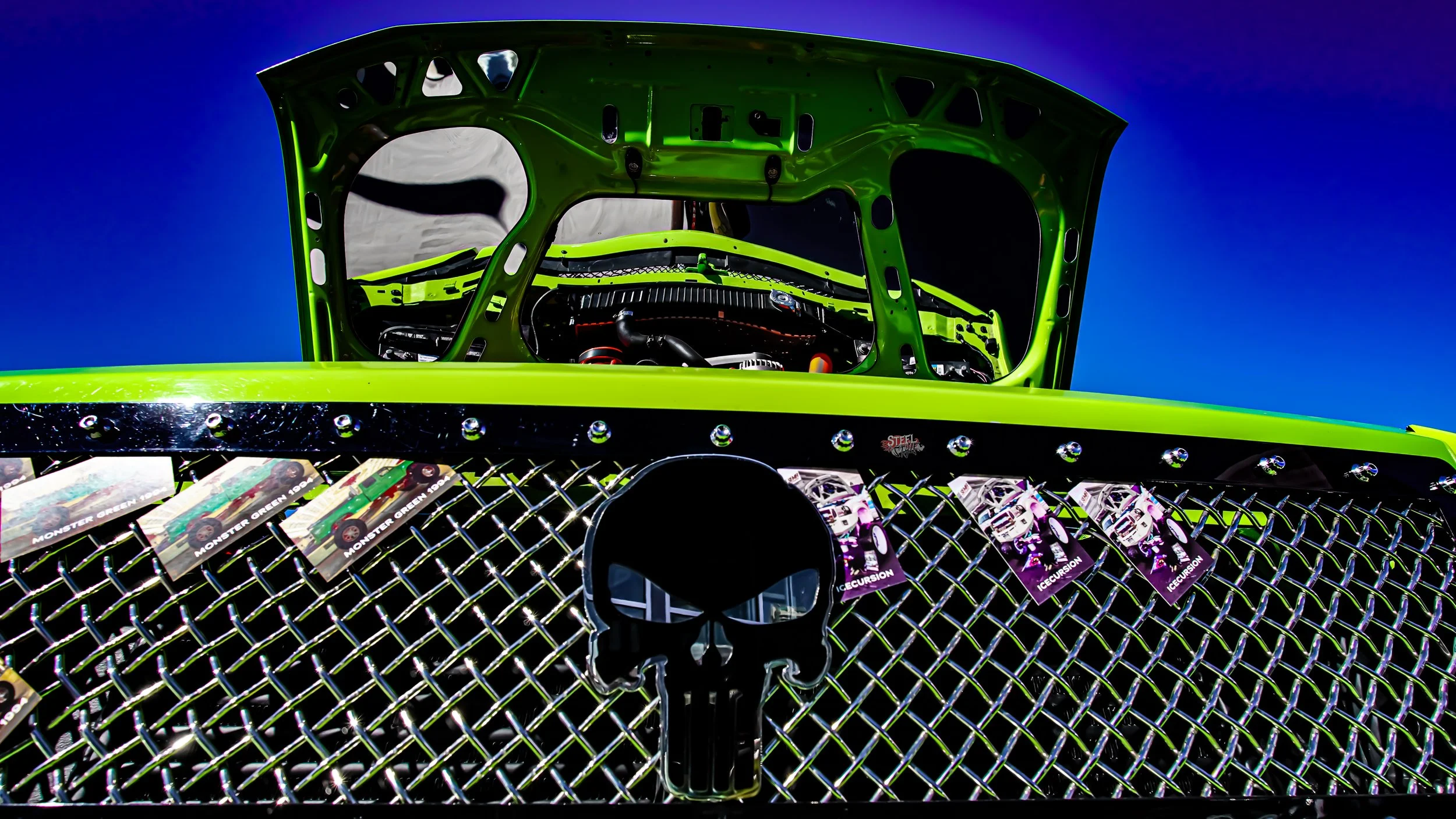 Close-up of a brightly colored race car with a green hood, skull emblem, and a diamond-patterned grille with stickers, under a bright blue sky.