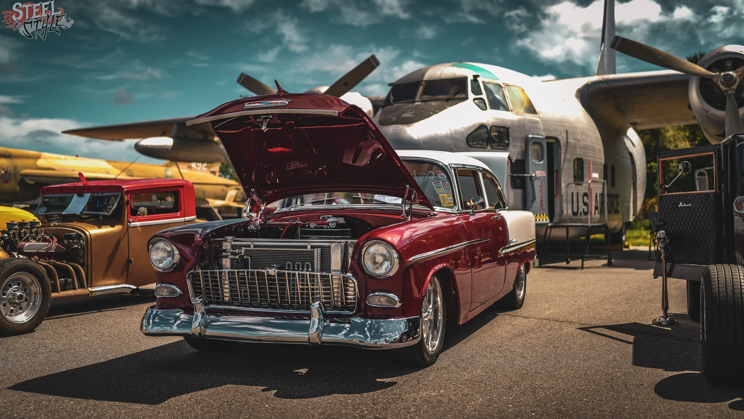 A vintage red car with its hood open displayed at an outdoor car show, with a large aircraft and various other old vehicles in the background under a partly cloudy sky.