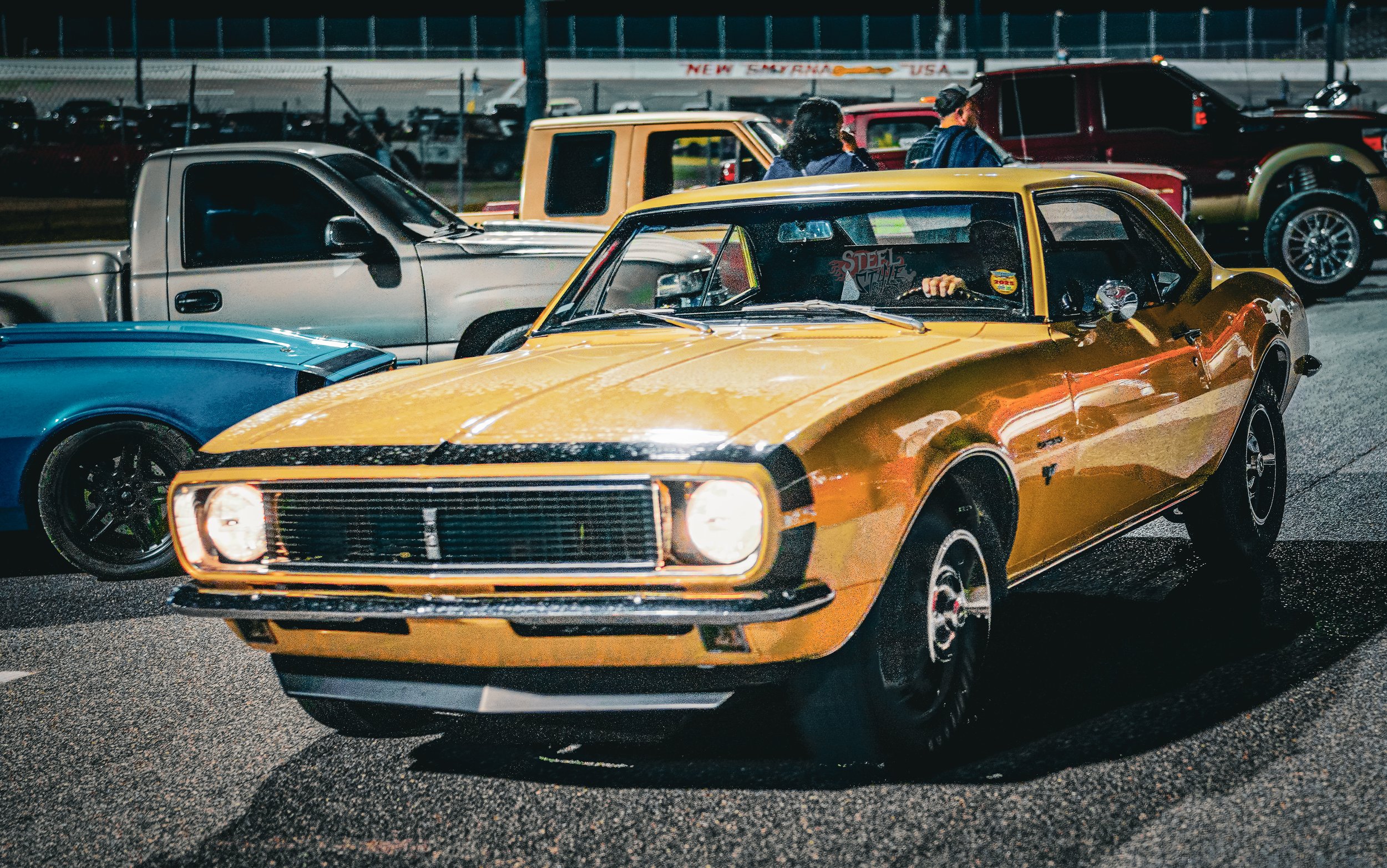 A vintage yellow muscle car parked at a car show at night, with other classic and modern trucks and cars around.