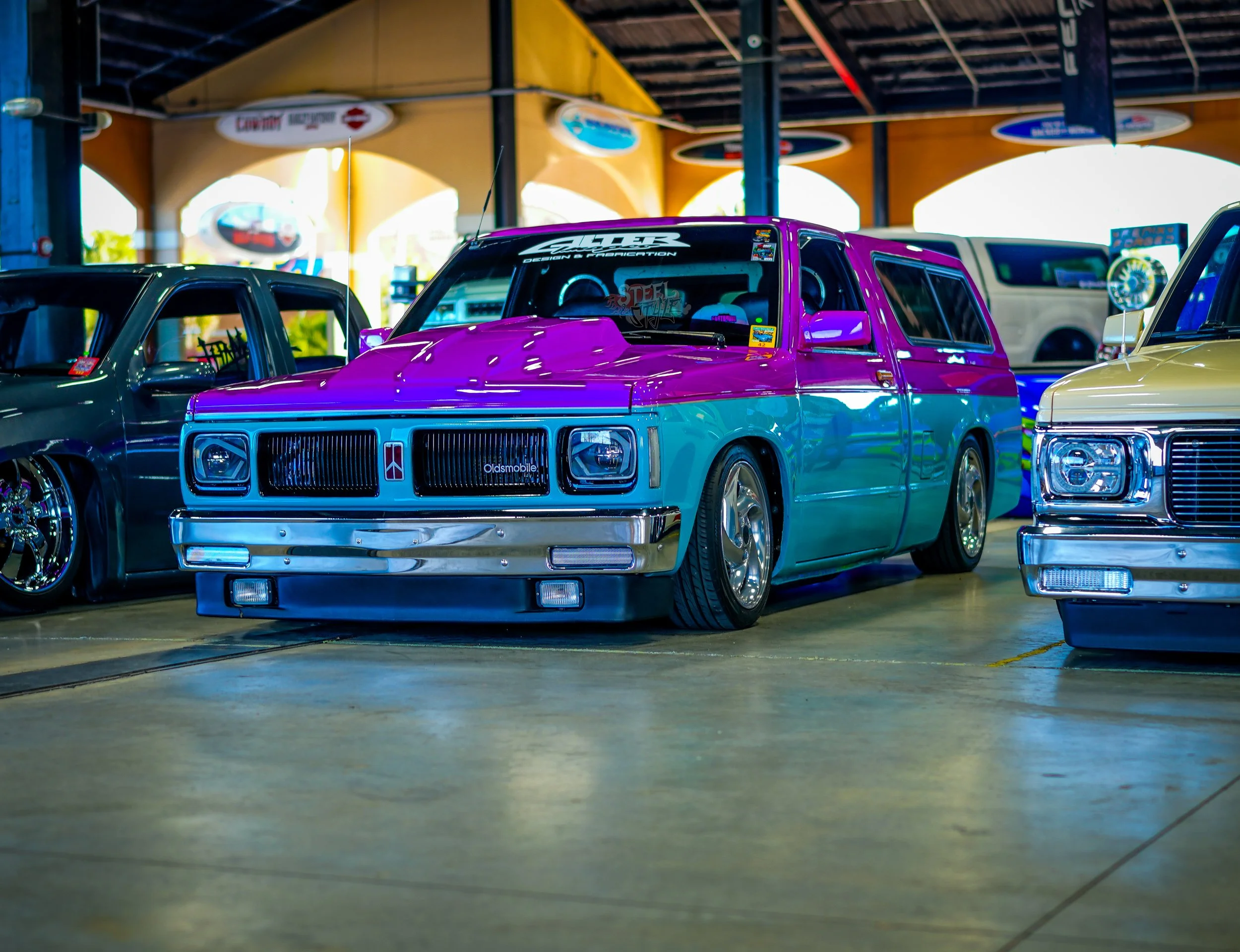 A vintage customized Oldsmobile car with a two-tone purple and teal paint job, lowered stance, and aftermarket wheels at a car show.