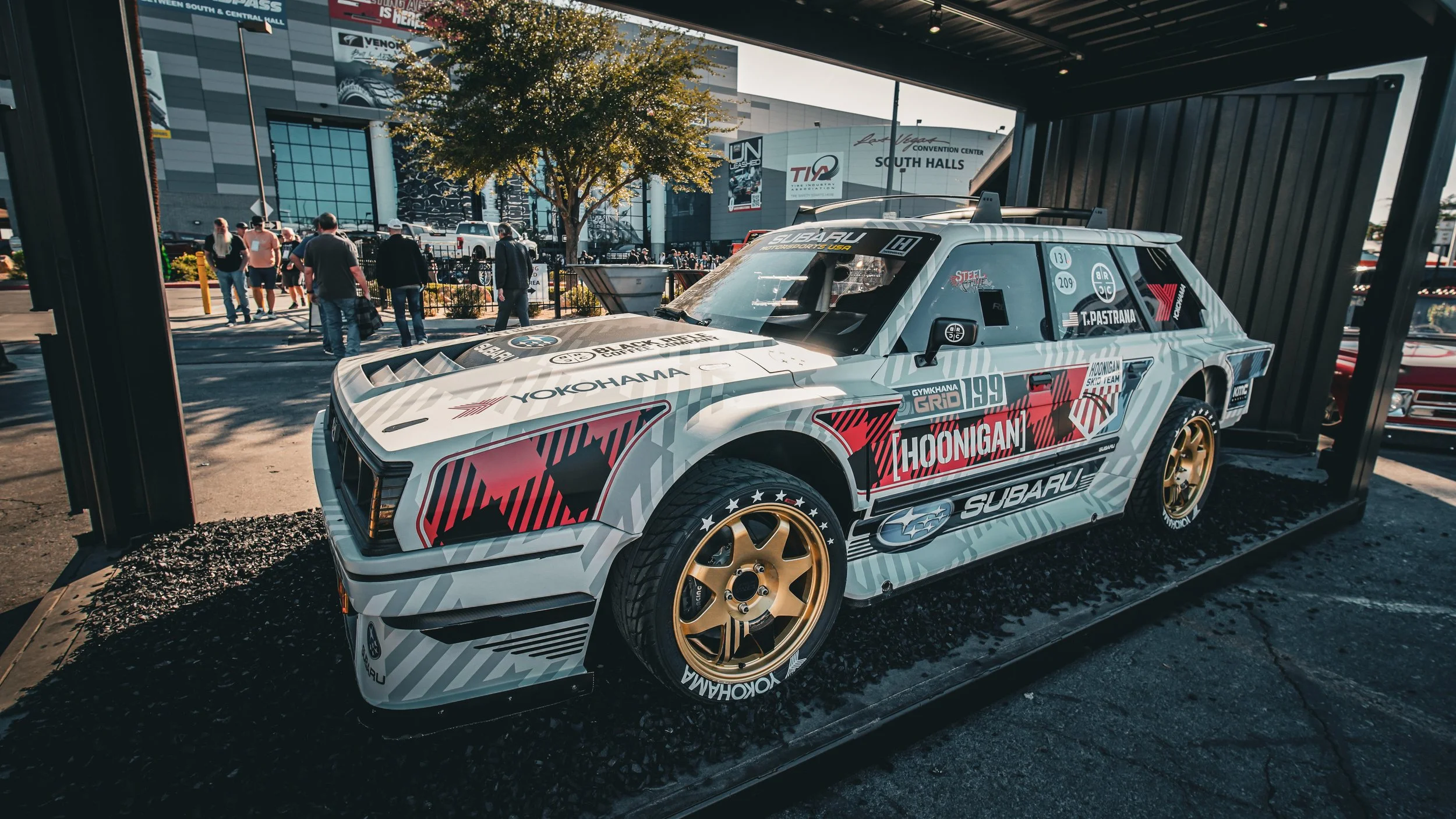 A vintage Subaru race car displayed outdoors under a shelter with a crowd and convention center in the background.