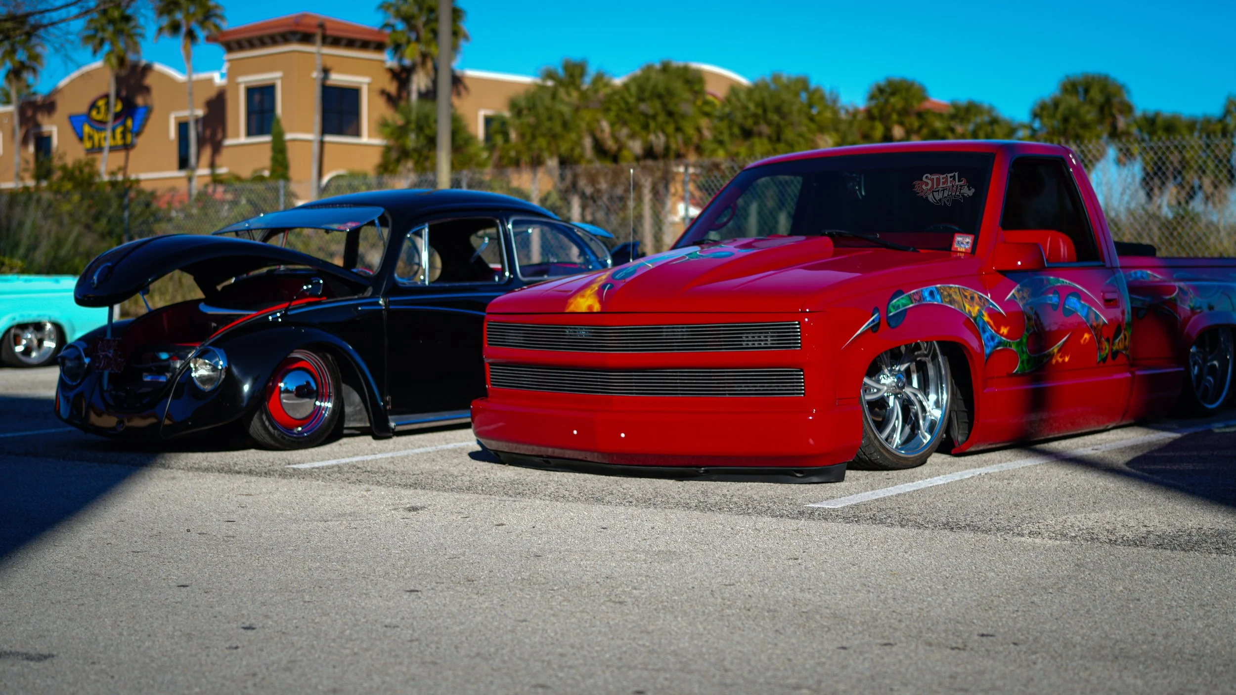 A black vintage Volkswagen Beetle with red hubcaps and an open engine hood, and a red custom lowrider pickup truck with colorful flame and dragon graphics on the sides, parked side by side in an outdoor parking lot with a chain-link fence and buildin