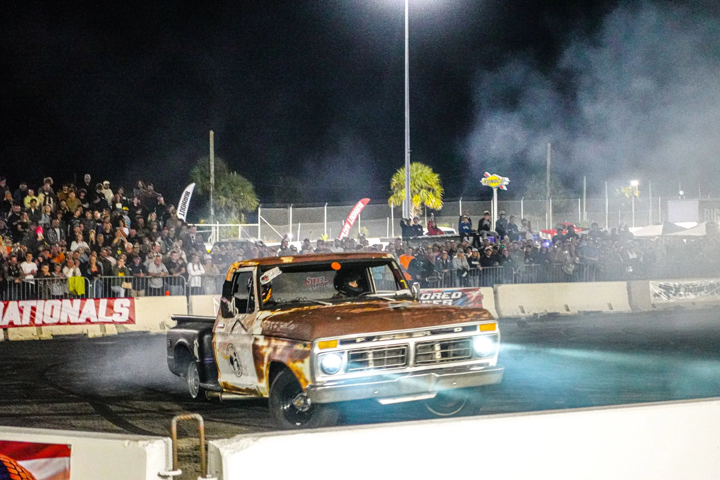 A vintage Ford pickup truck with a rusted exterior driving on a dirt track at night during a racing event, with a large crowd of spectators behind a fence watching.