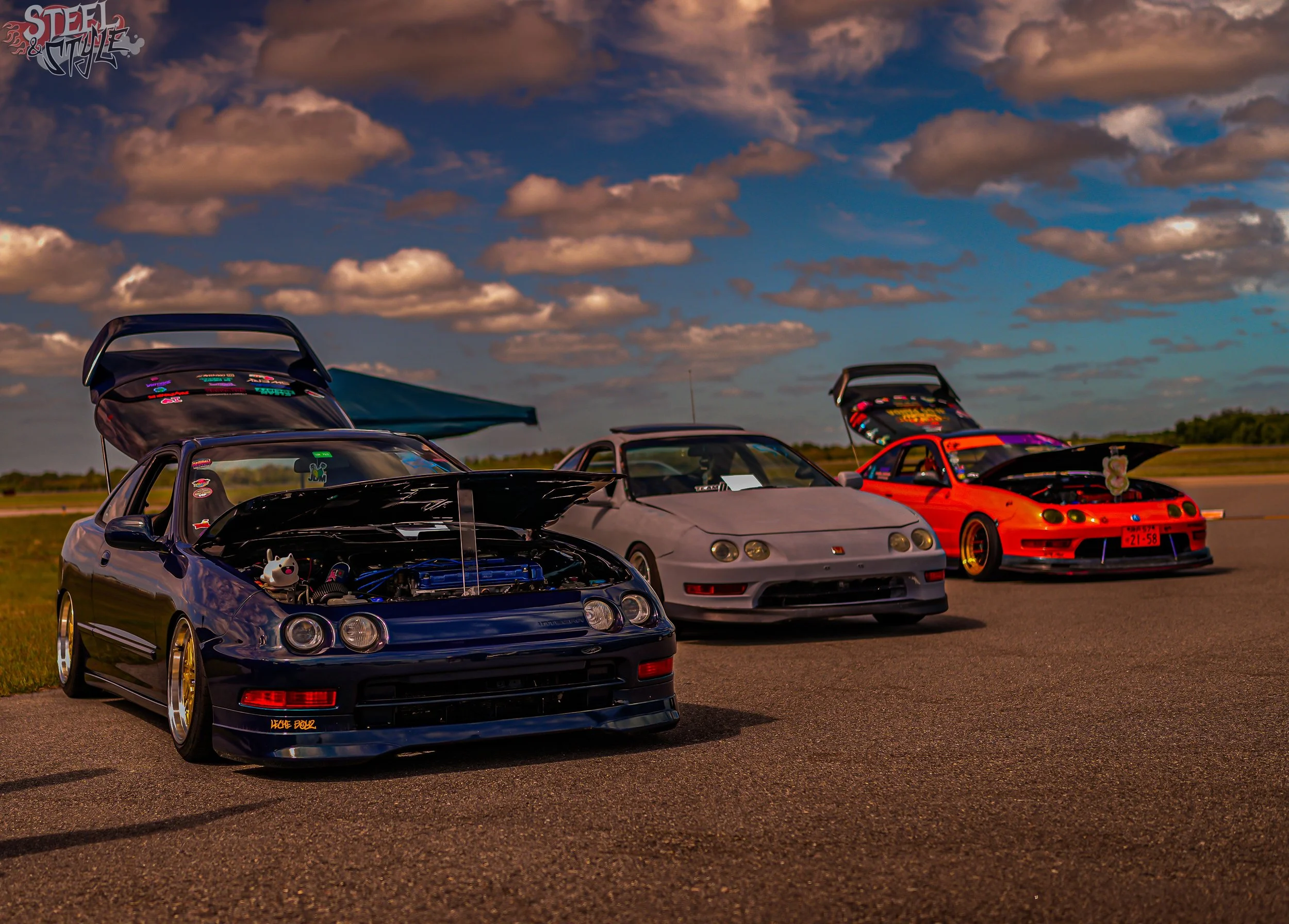 A lineup of four sports cars with their hoods open, parked outdoors under a cloudy sky during the day, displaying various customizations and stickers.
