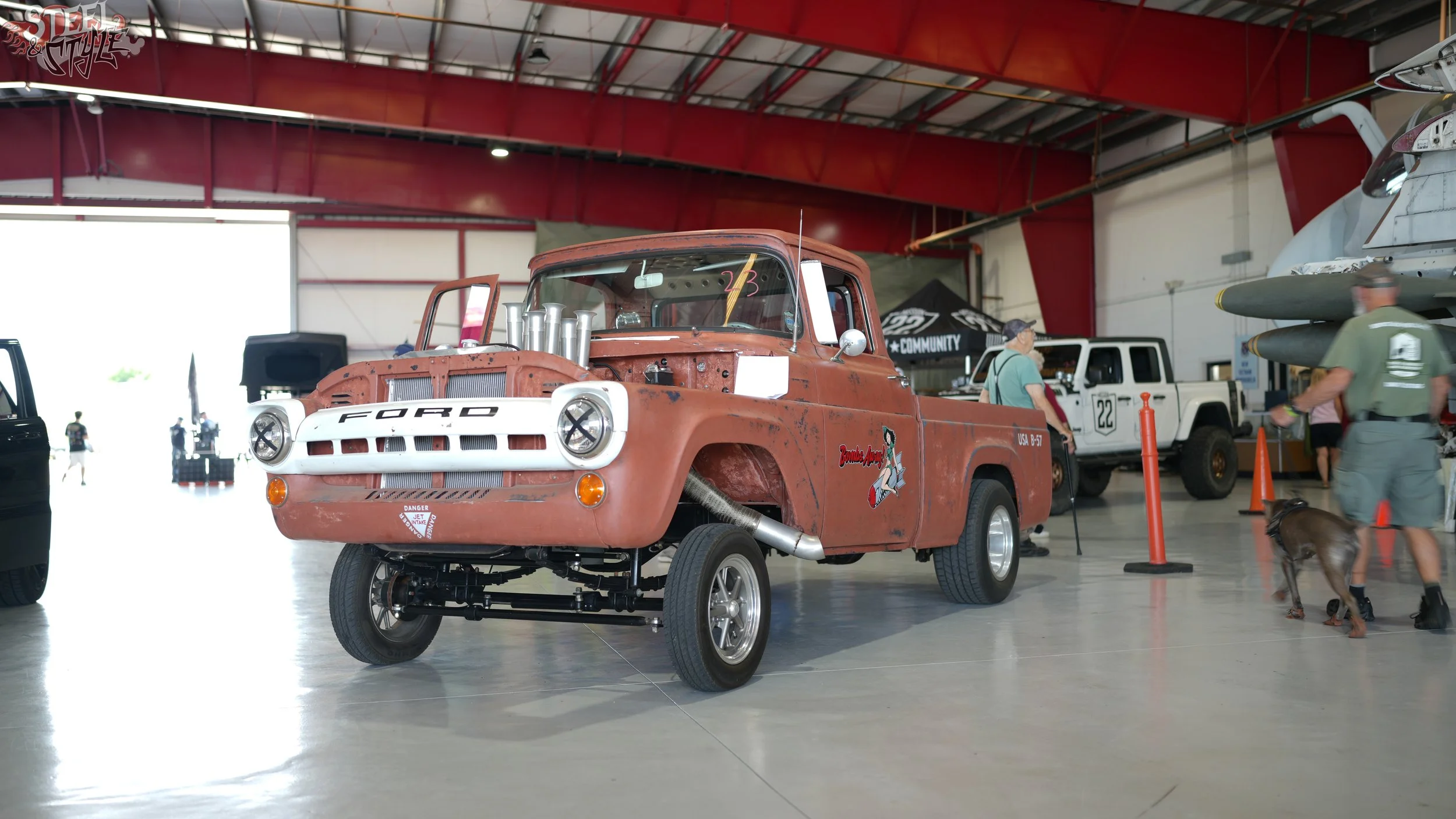 A rusty vintage pickup truck with exposed engine parts on display at an indoor car show, with other vehicles and people in the background.