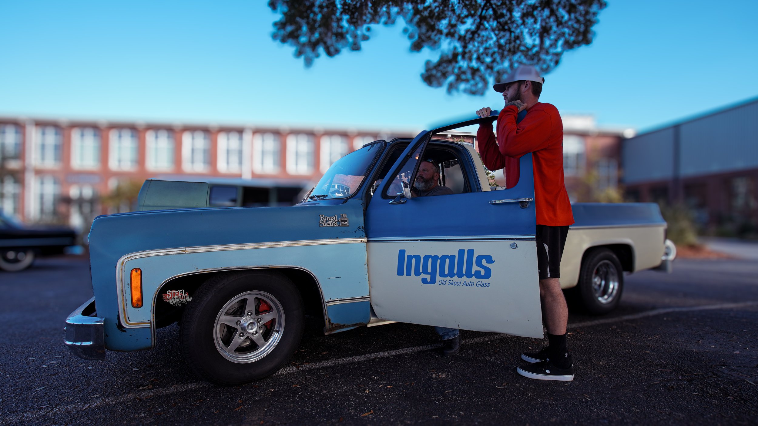 A man in a red jacket, black shorts, and sneakers stands outside a vintage blue pickup truck, talking to the driver inside. The truck has a sign for Ingalls Auto Glass on the door and is parked in a lot with a building and another vehicle in the back