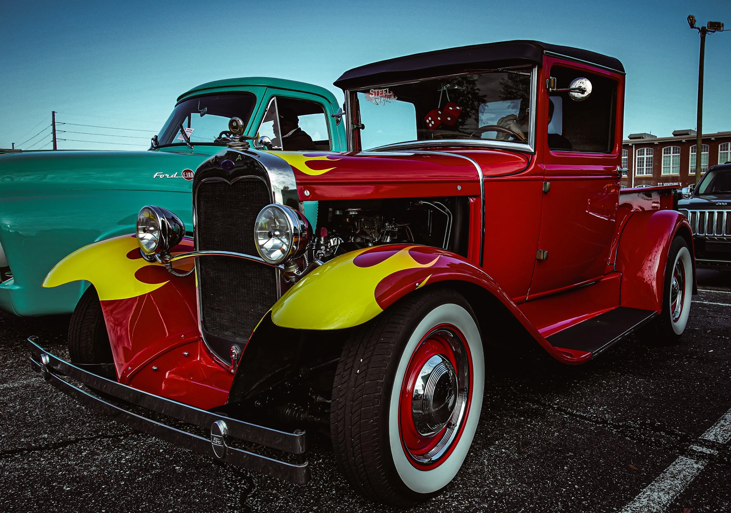 A vintage red and yellow hot rod car with a flame design parked in a lot, with a teal classic car and other vehicles in the background.