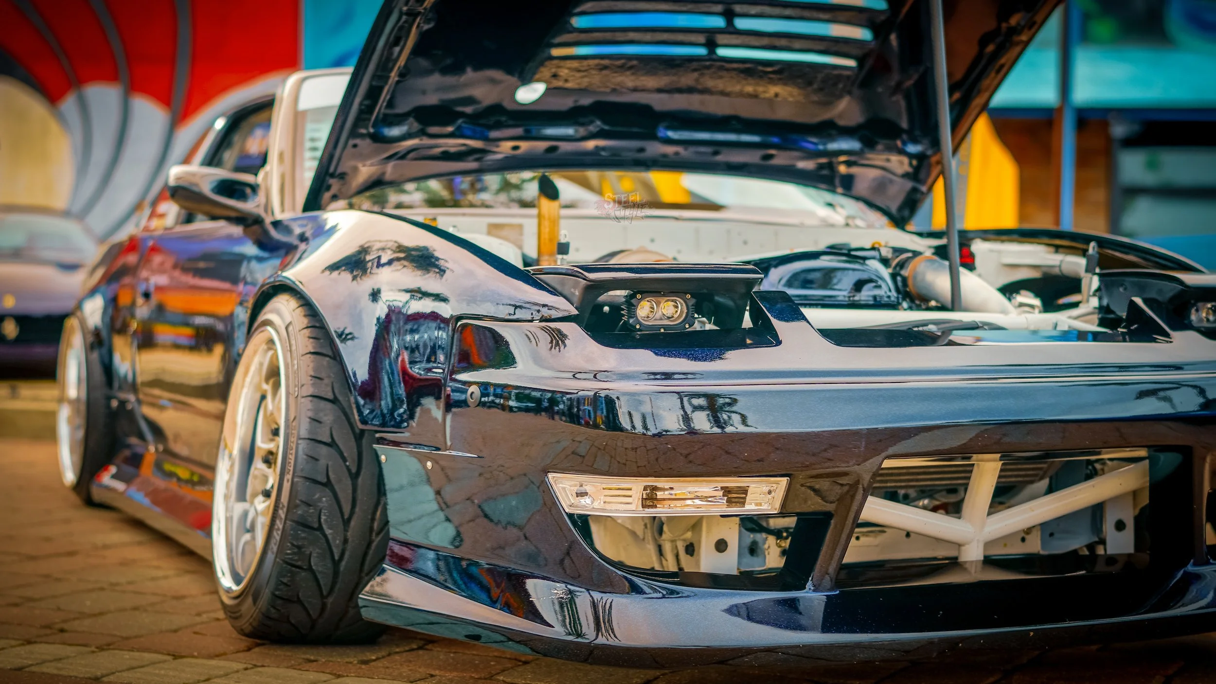 A sleek black sports car with its hood open, revealing the engine, parked on a brick surface at a car show. The car has wide tires and a reflective surface.