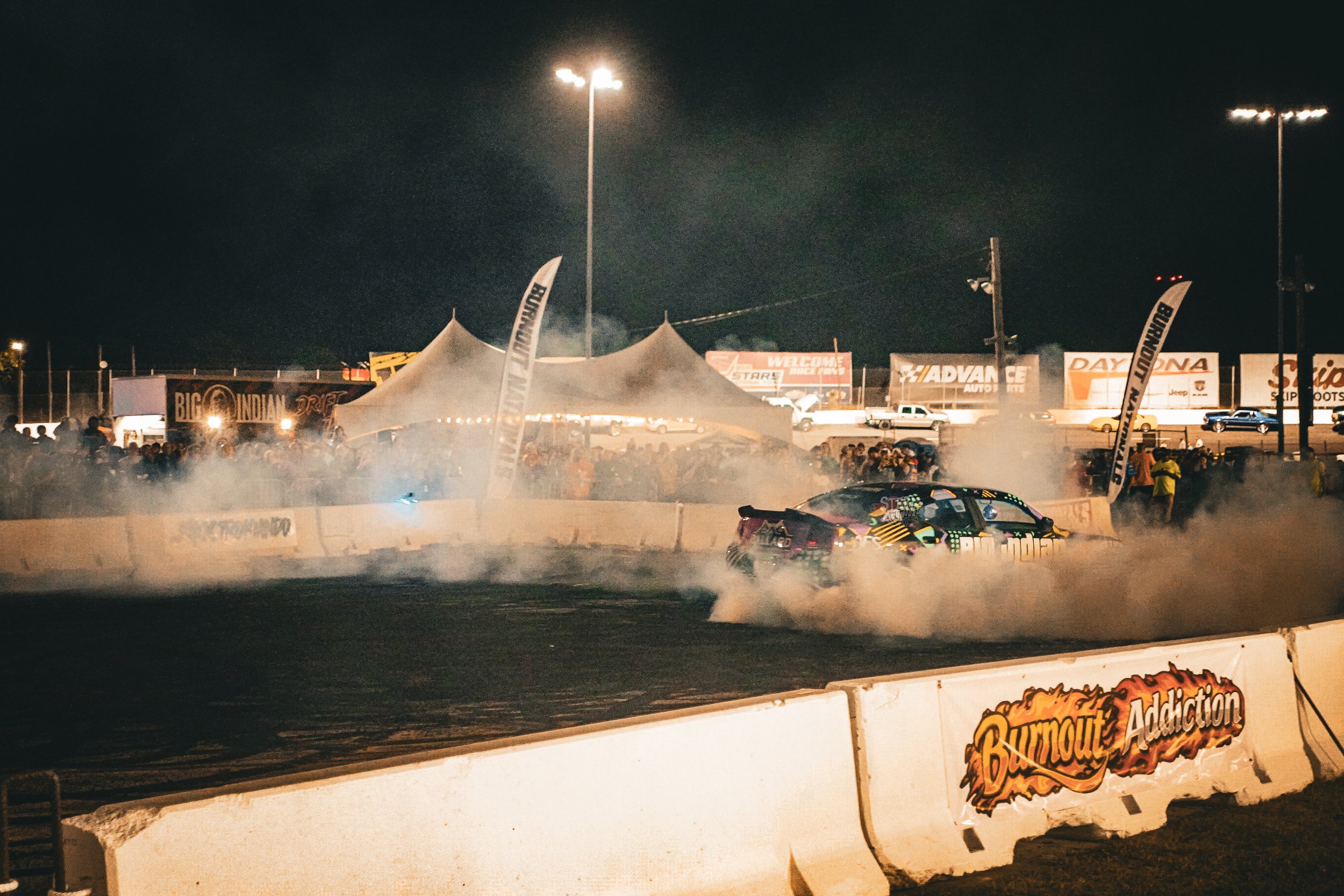 A race car drifting on a dirt track at night, with a large crowd gathered behind barriers, tents, and banners around the track. Smoke and dust are visible from the car's tires, with bright stadium lights overhead.