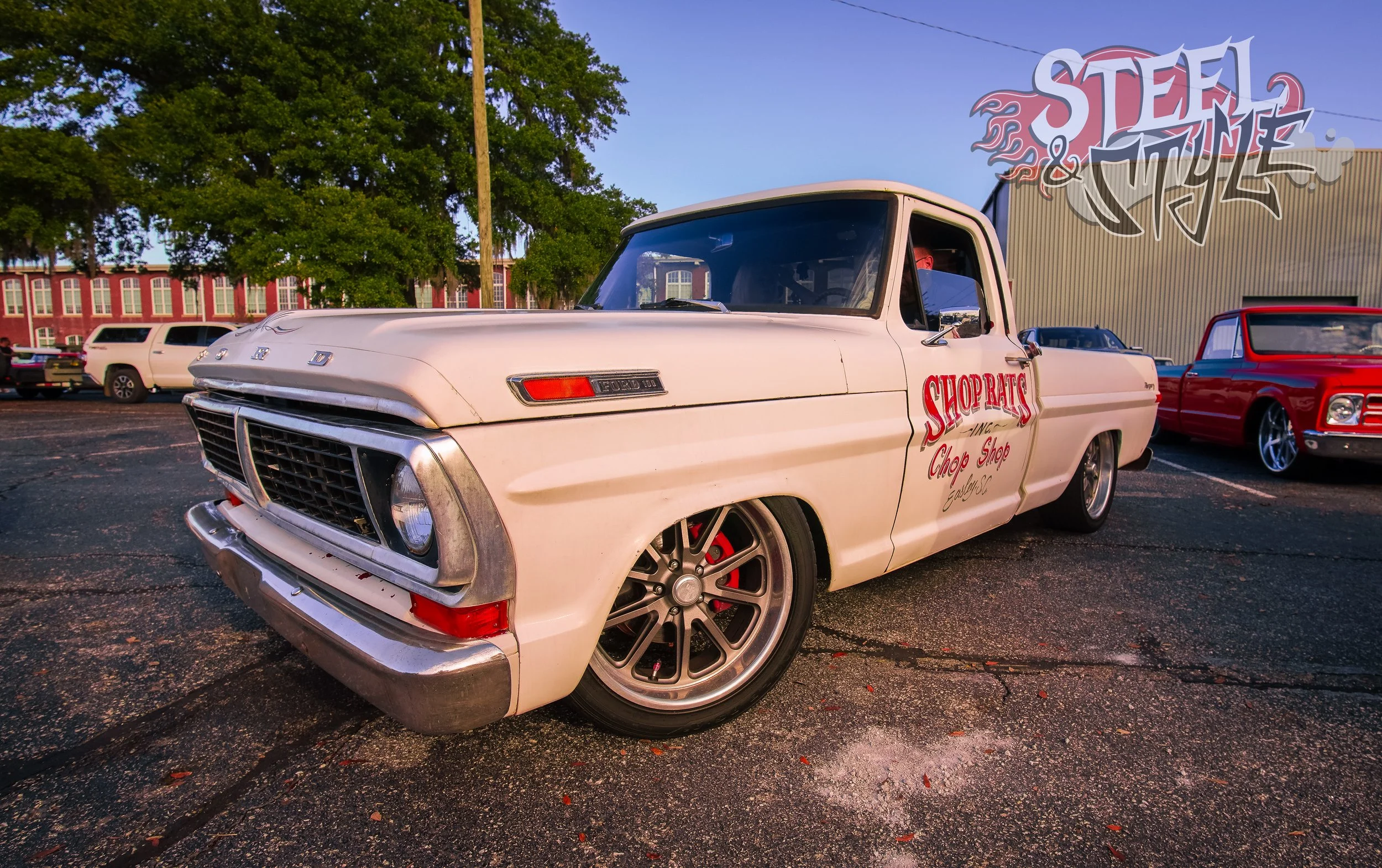A vintage white pickup truck parked in a lot with a sign on its door for Shop Ratz game chop shop. Other classic cars are visible in the background, along with trees and a building. The photo is taken during daytime.