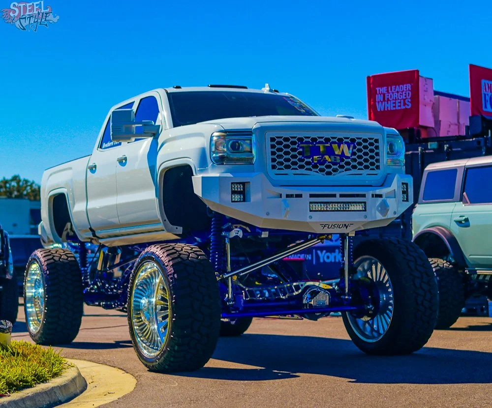 A white lifted truck with large wheels and tires, custom suspension, and a front grille with 'TTW' and 'FUSION' branding, parked outdoors in a parking lot.