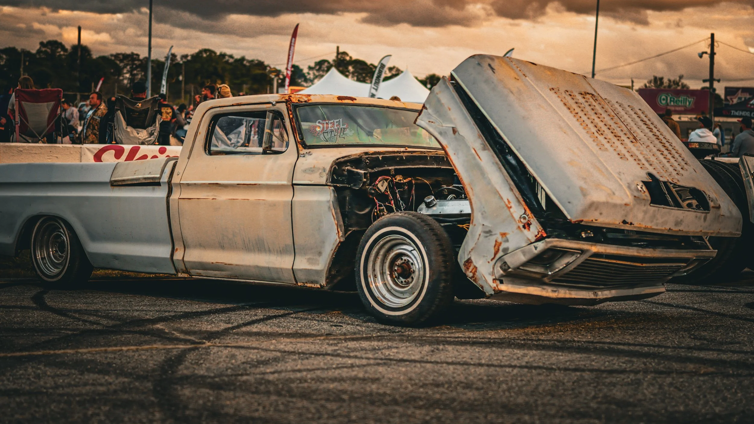 A rusty, battered vintage pickup truck with its hood open at a car show, with people and tents in the background.