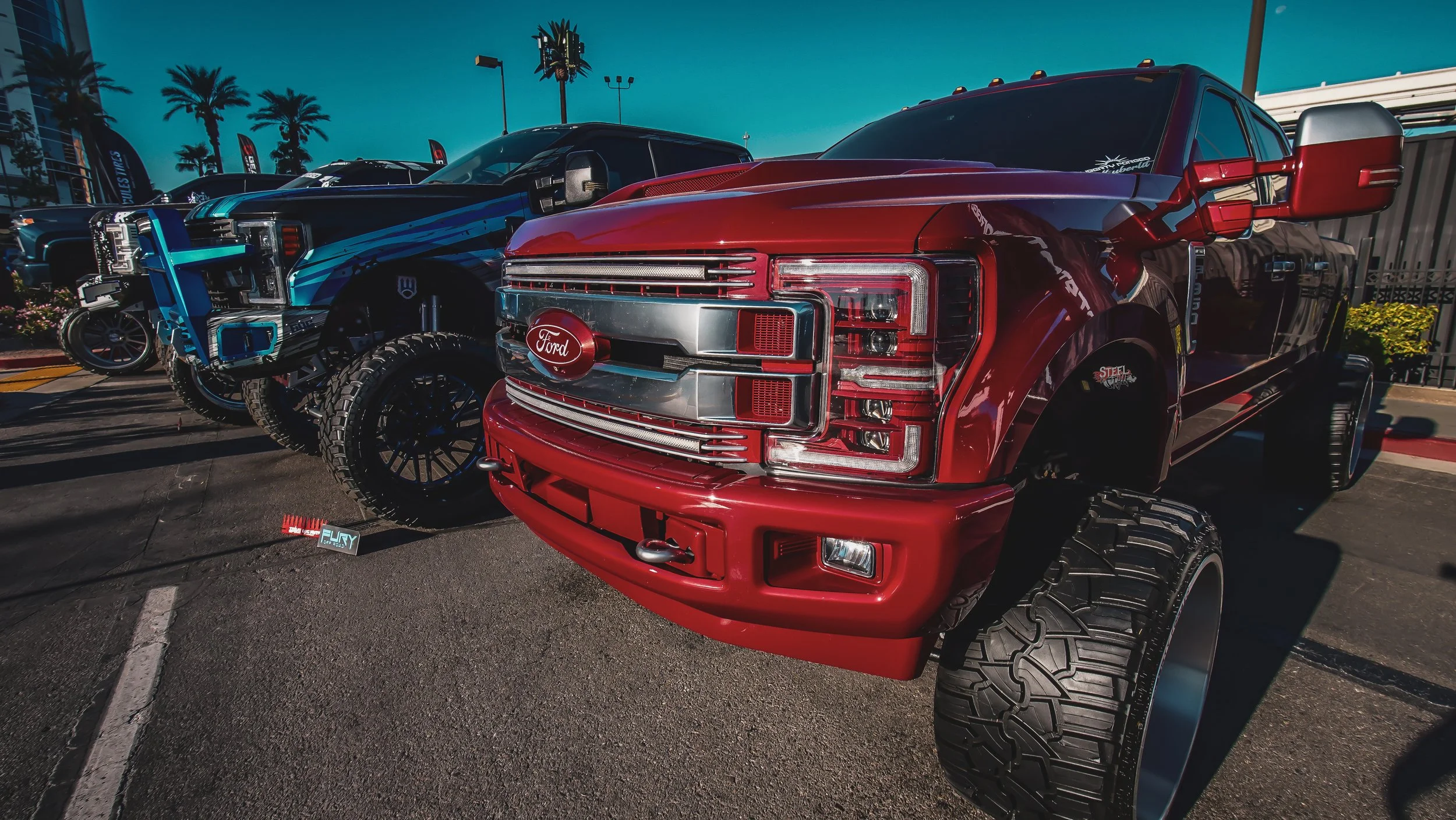 Red Ford truck with oversized tires parked outdoors with other vehicles and palm trees visible in the background.