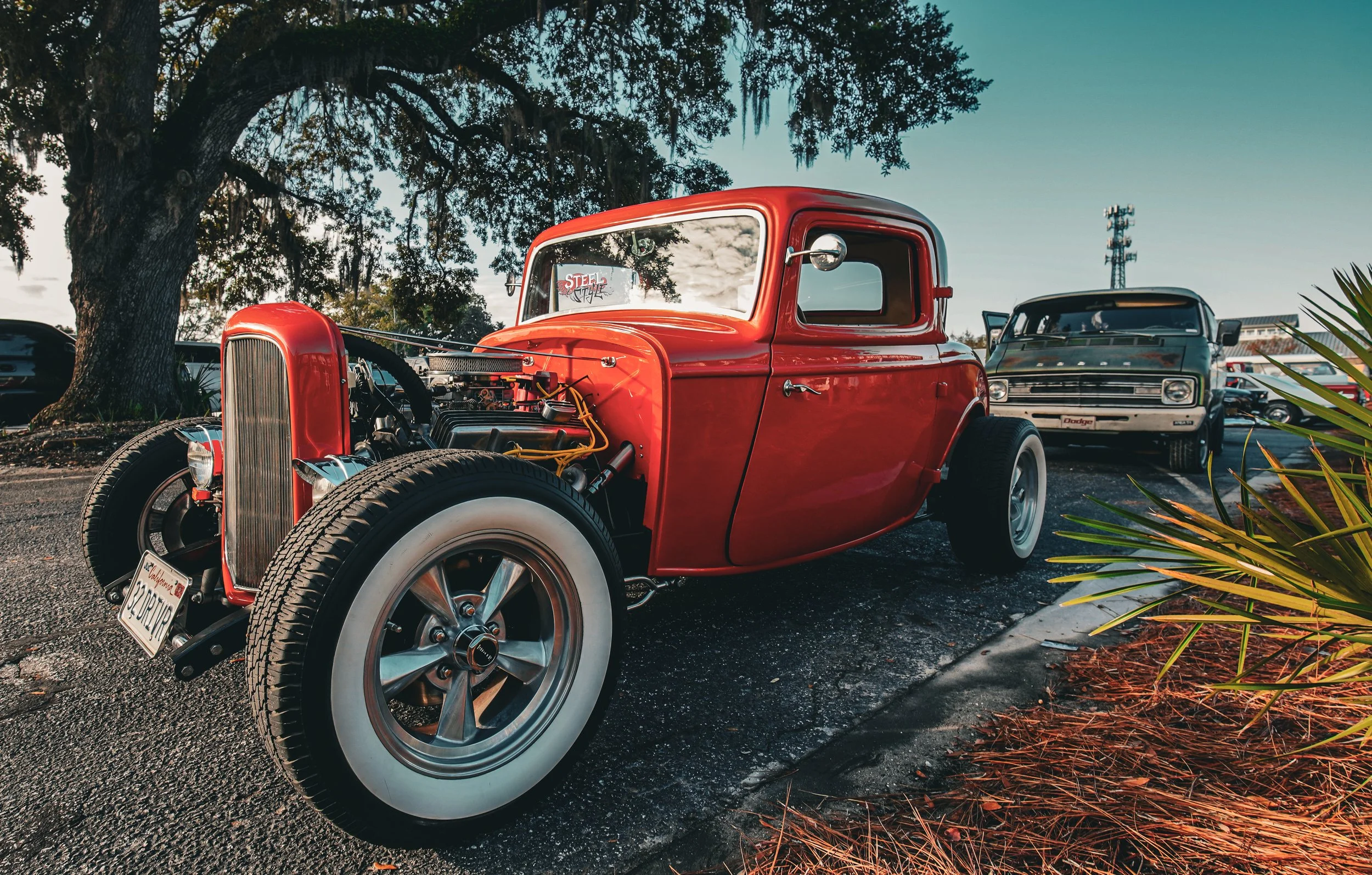 Red vintage hot rod car with exposed engine parked on asphalt, with a large tree and a black van in the background, and plants with reddish leaves in the foreground.