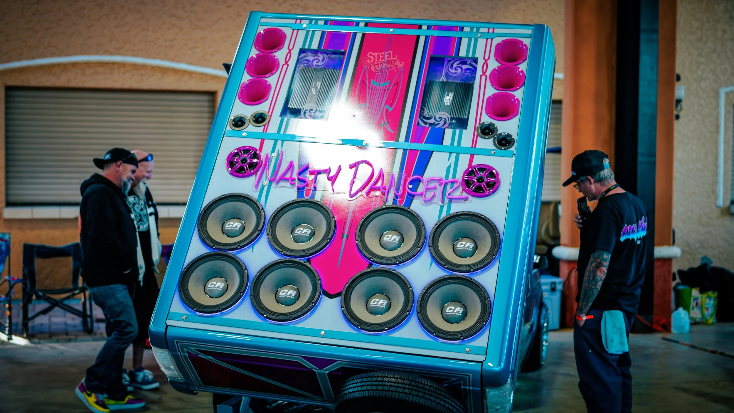 A large, colorful jukebox with a neon sign that reads 'NASTY DANCER' displayed on it, surrounded by three people in a room.