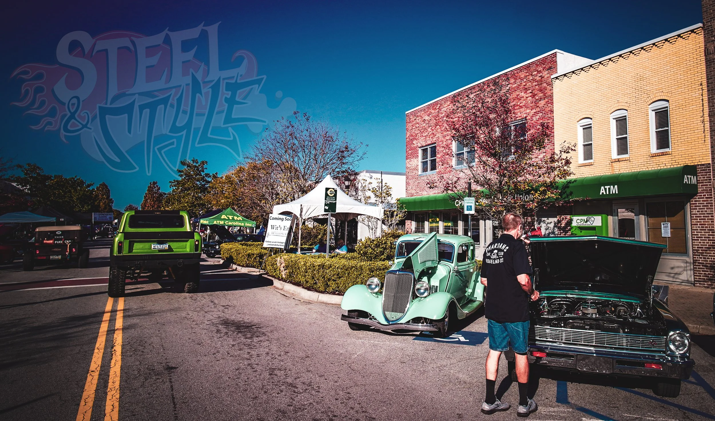 A street scene featuring vintage cars parked in front of a brick building with a green ATM. A man is inspecting the open hood of a black car. There are tents and signs indicating a community event or car show, with trees lining the street under a blu