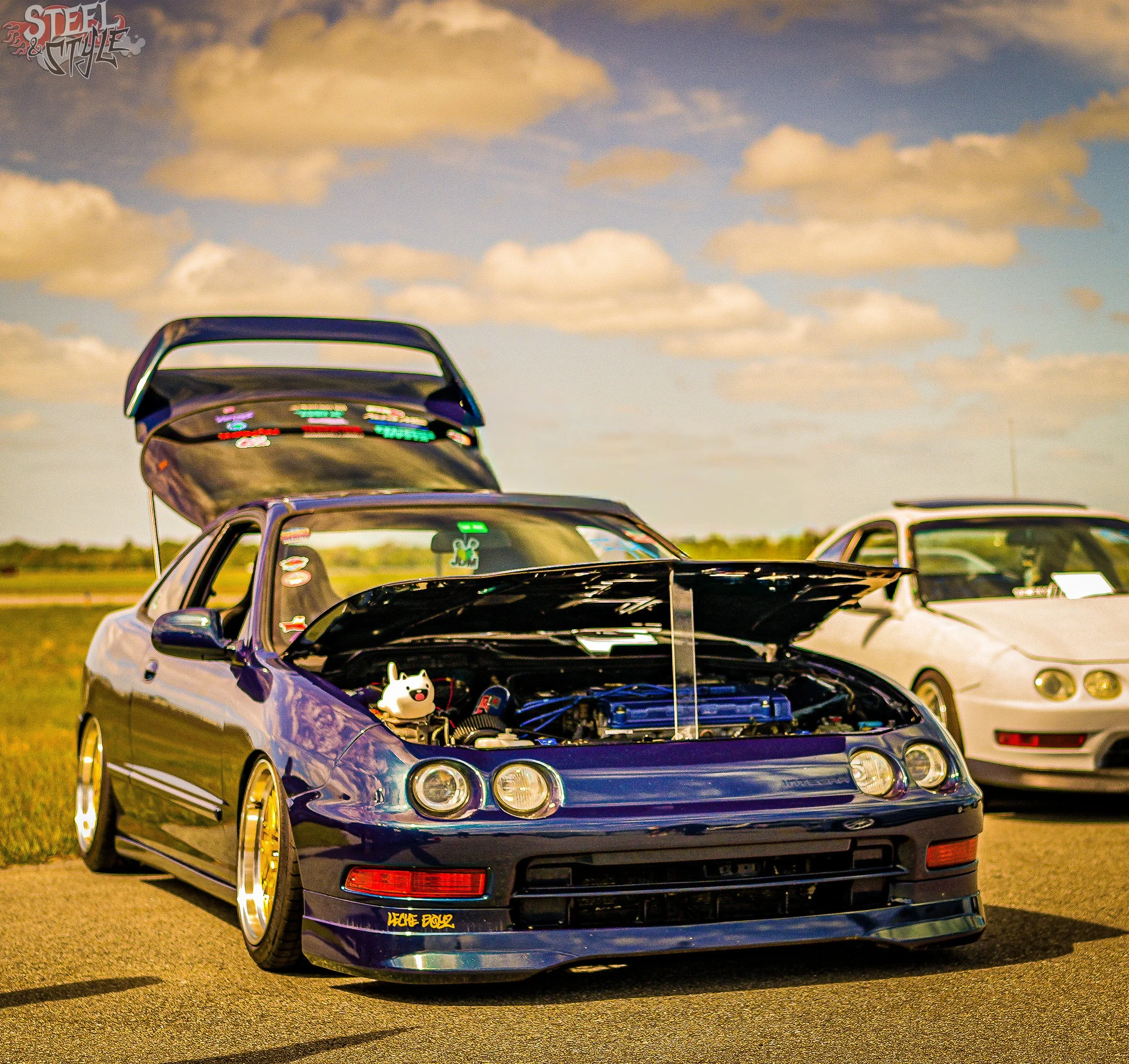 A dark-colored sports car with its hood open, parked on an open lot during daytime with scattered clouds in the sky.