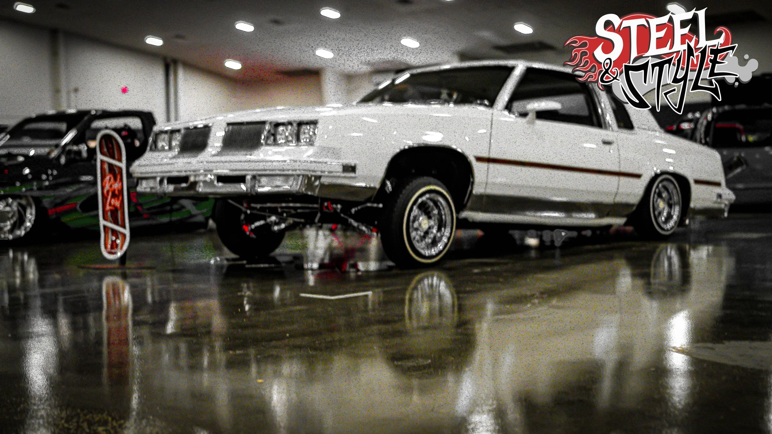 A classic white luxury car on display at an indoor car show, with a 'Steel and Style' logo in the top right corner.