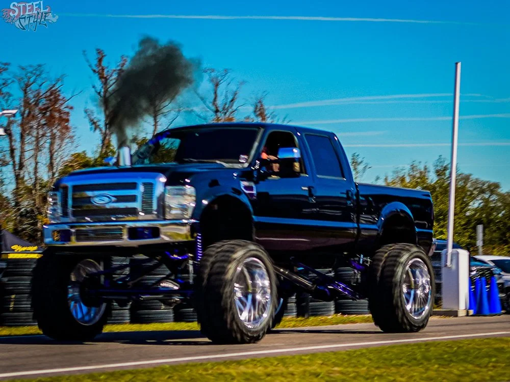 A modified black pickup truck with oversized wheels and lift kit, emitting black smoke from the exhaust, driving on a race track during the day.