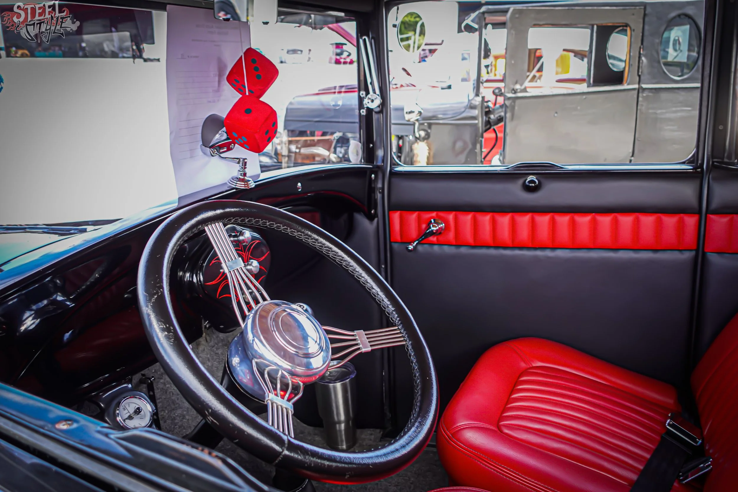 Inside the driver’s area of a classic car with a black and red interior. The dashboard features a chrome steering wheel, a gauge, and an exposed wiring connection. There are two red dice hanging from the rearview mirror and a red leather seat on the 