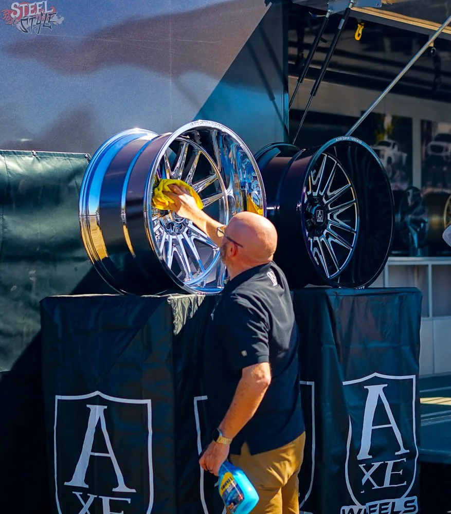 A man cleaning shiny, chrome car wheels at an outdoor event, with two large wheels displayed on a table covered with a black cloth featuring a logo.