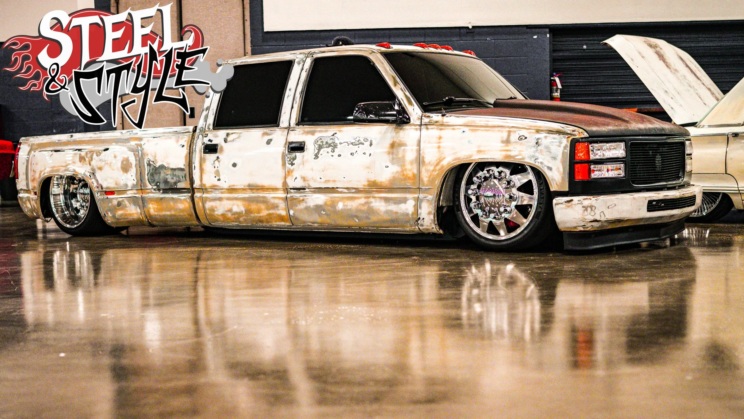A heavily modified, rusty, lowered pickup truck with custom chrome wheels at an indoor car show, with a graffiti-style "Steel & Style" logo in the top left corner.