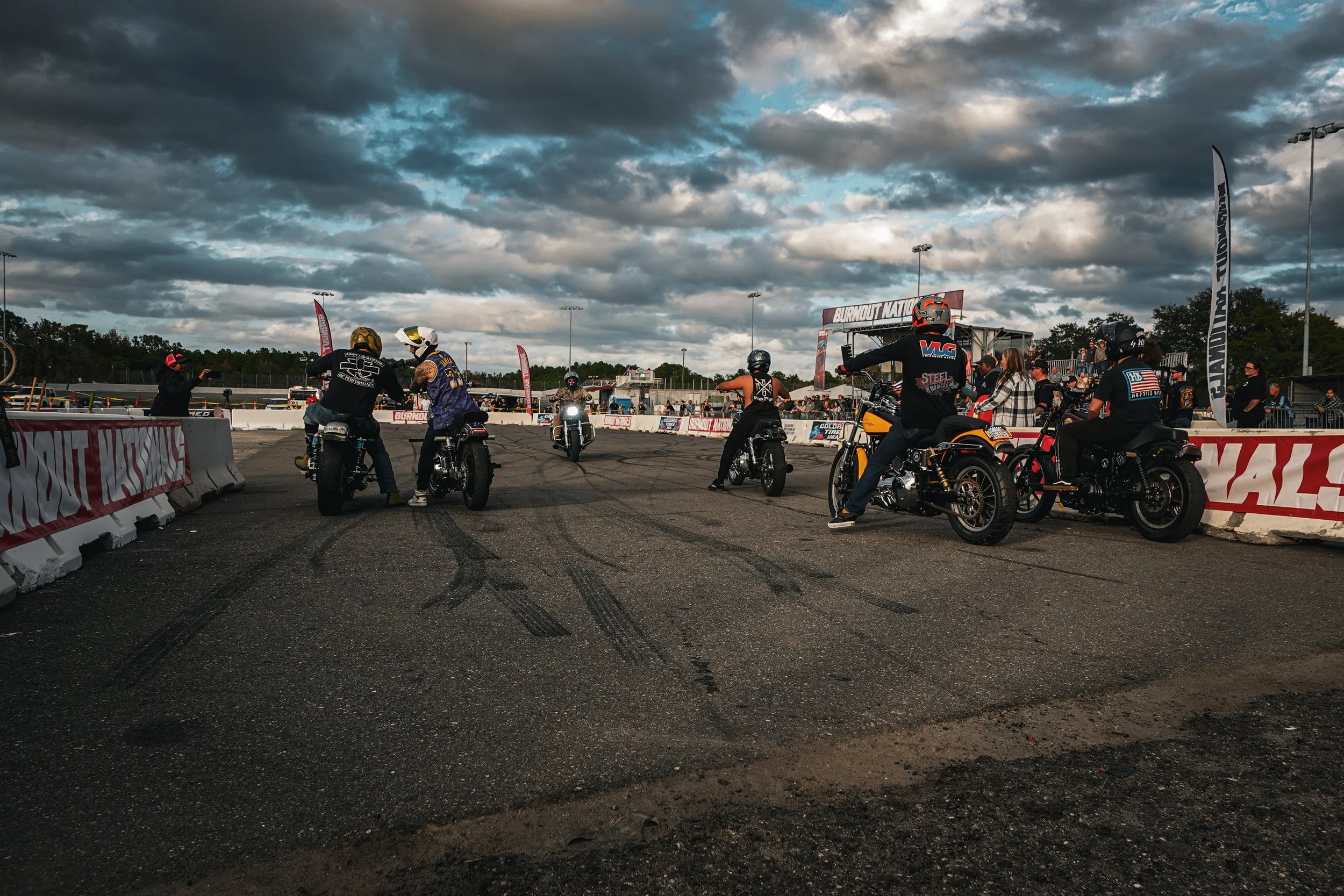 Motorcycles and riders gathered at a racetrack, with some taking photos, under cloudy skies.