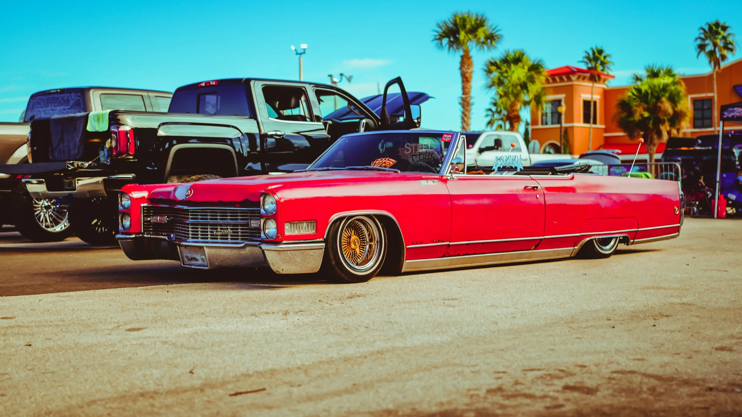 A vintage red Cadillac lowered on the ground at a car show, with other vehicles and palm trees in the background under a blue sky.