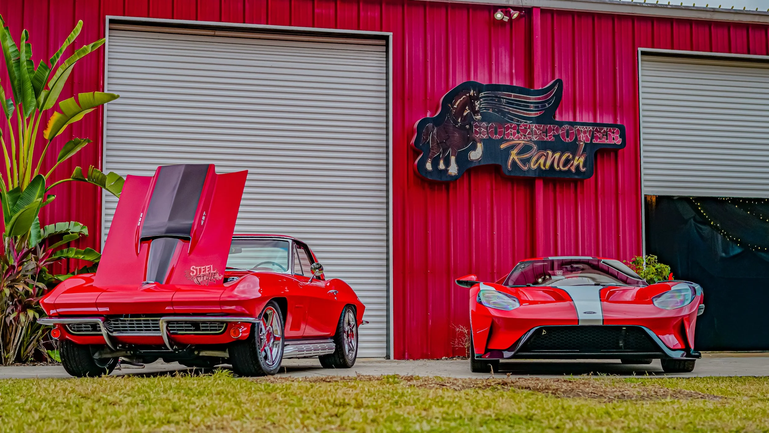 Two red sports cars parked in front of a red building with a sign reading 'Horsepower Ranch.' The car on the left is a vintage model with its hood open, revealing the engine, and has a sticker that says 'Steel Stallion.' The car on the right is a mod