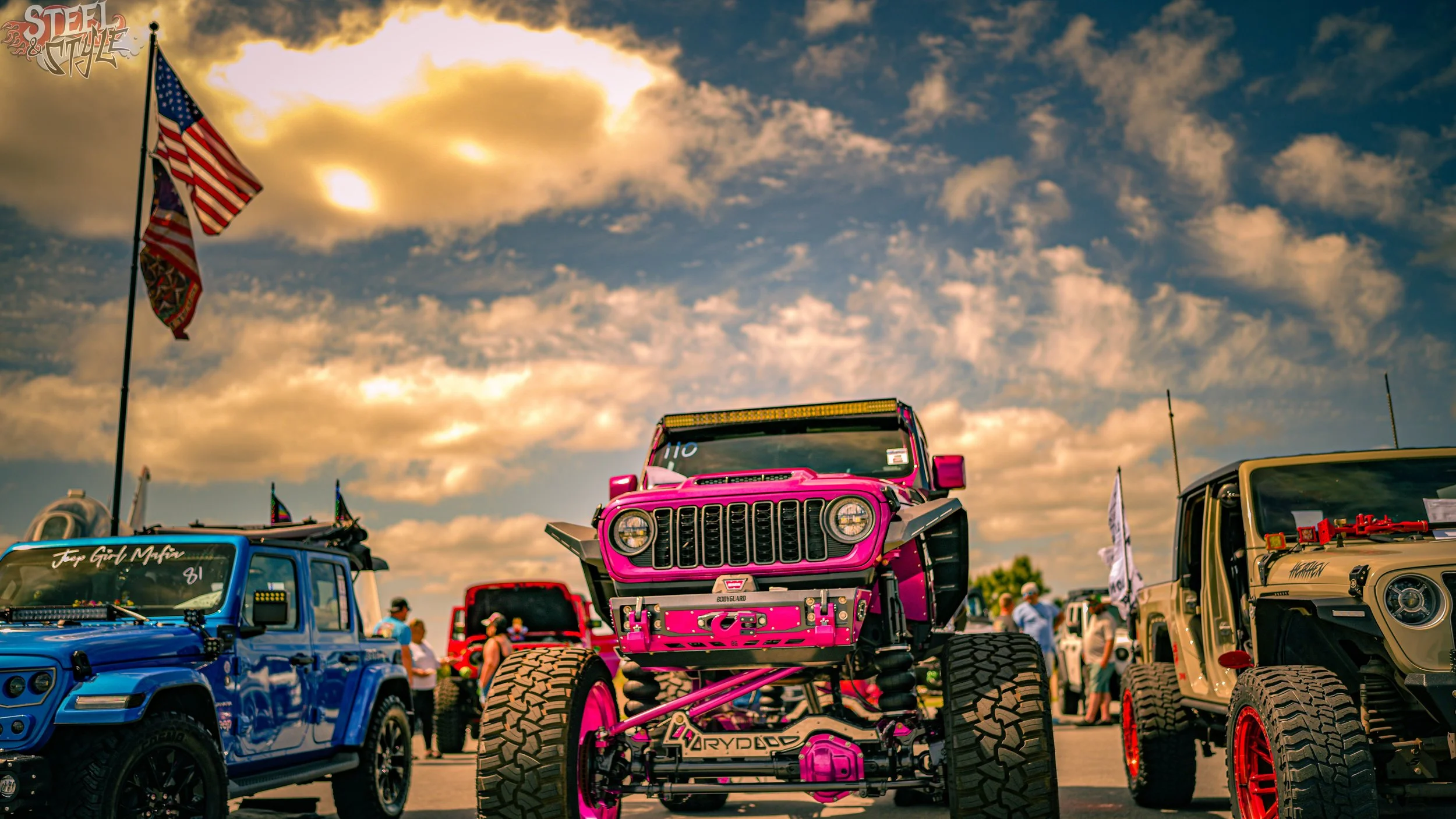 A bright pink monster truck displayed at an outdoor vehicle show, surrounded by other off-road vehicles and people under a partly cloudy sky.