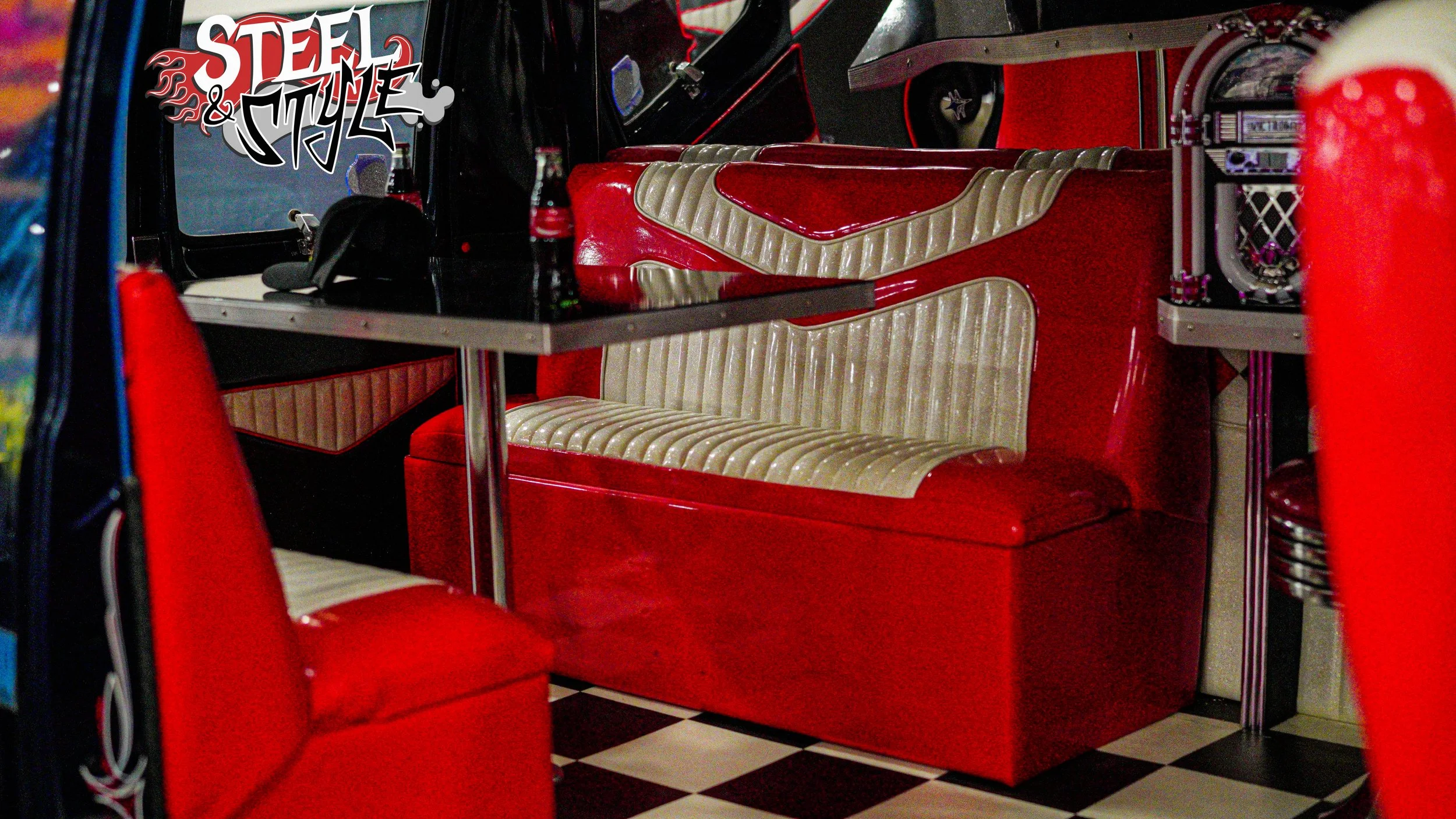 Vintage diner booth with red and cream vinyl seats and checkered floor, decorated with 'Steel & Style' sign, drinks, and a jukebox visible.