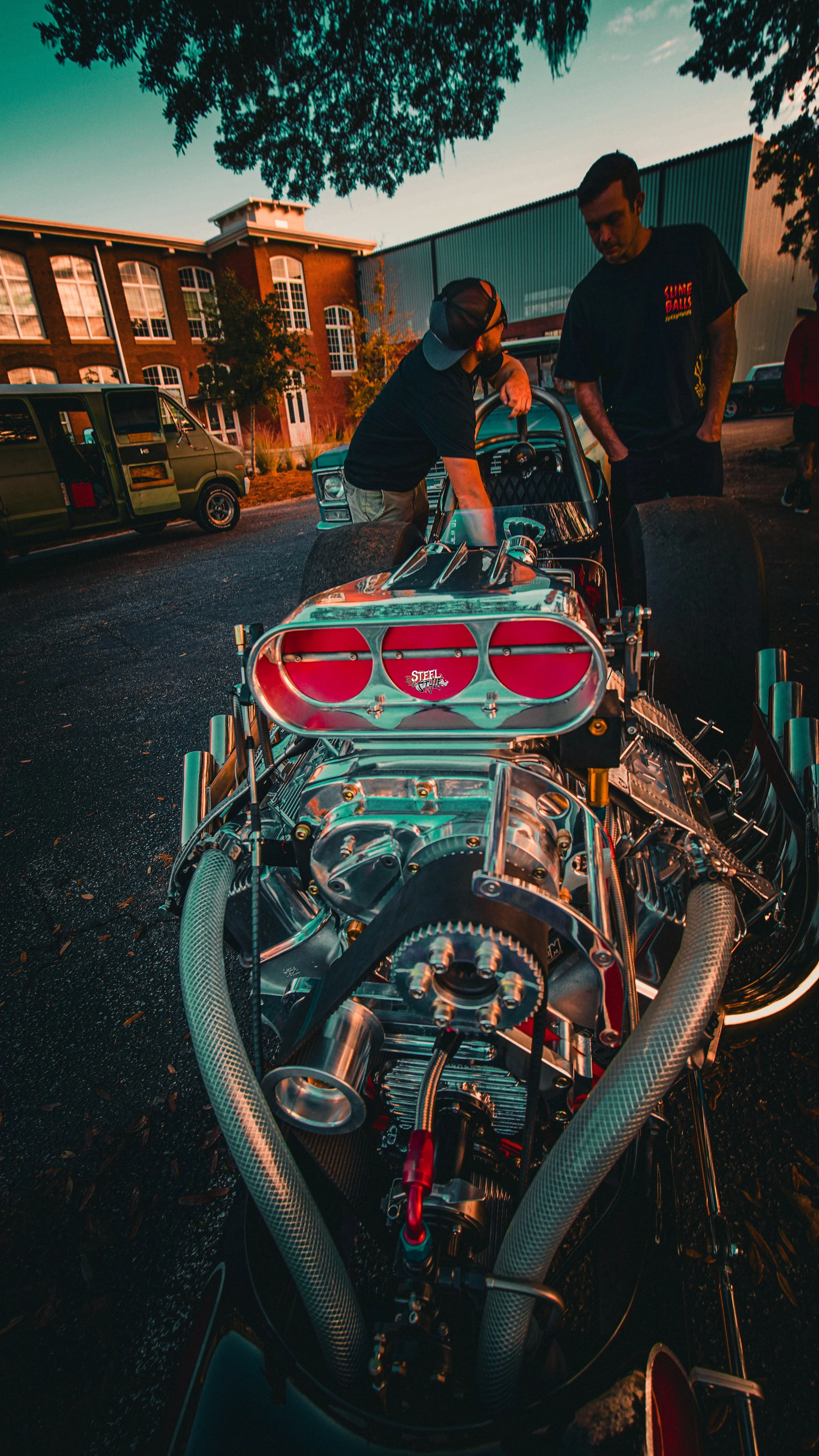 Two men are gathered around a vintage drag racing car with a polished engine, parked outdoors during sunset. The setting includes trees, a brick building, and other vehicles in the background.