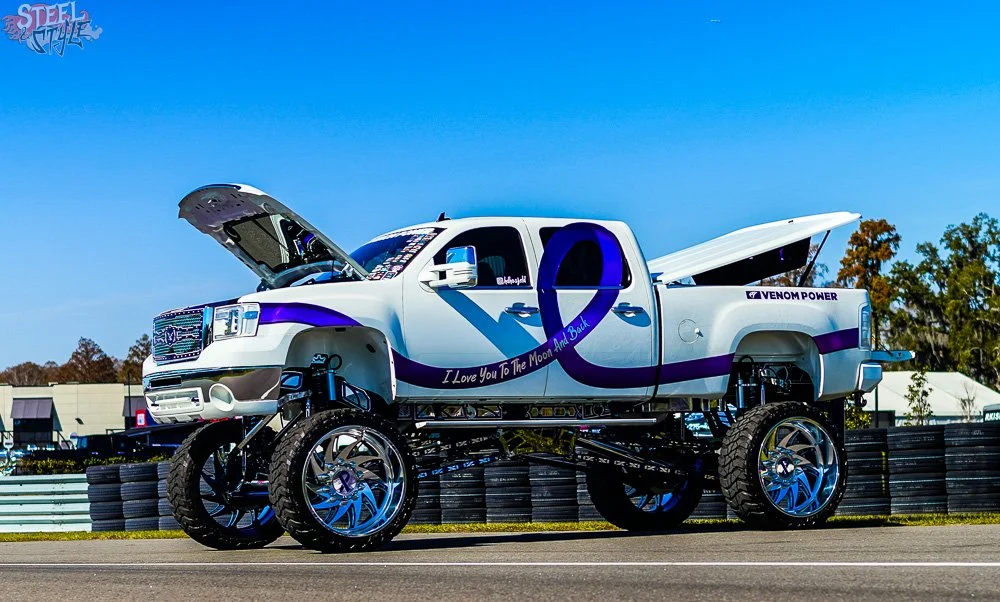 A white pickup truck with purple accents and graphics, including a purple ribbon and messages like "I Love You To The Moon And Back" and "Venom Power," modified with large off-road tires and suspension, is on a race track with a clear blue sky and so