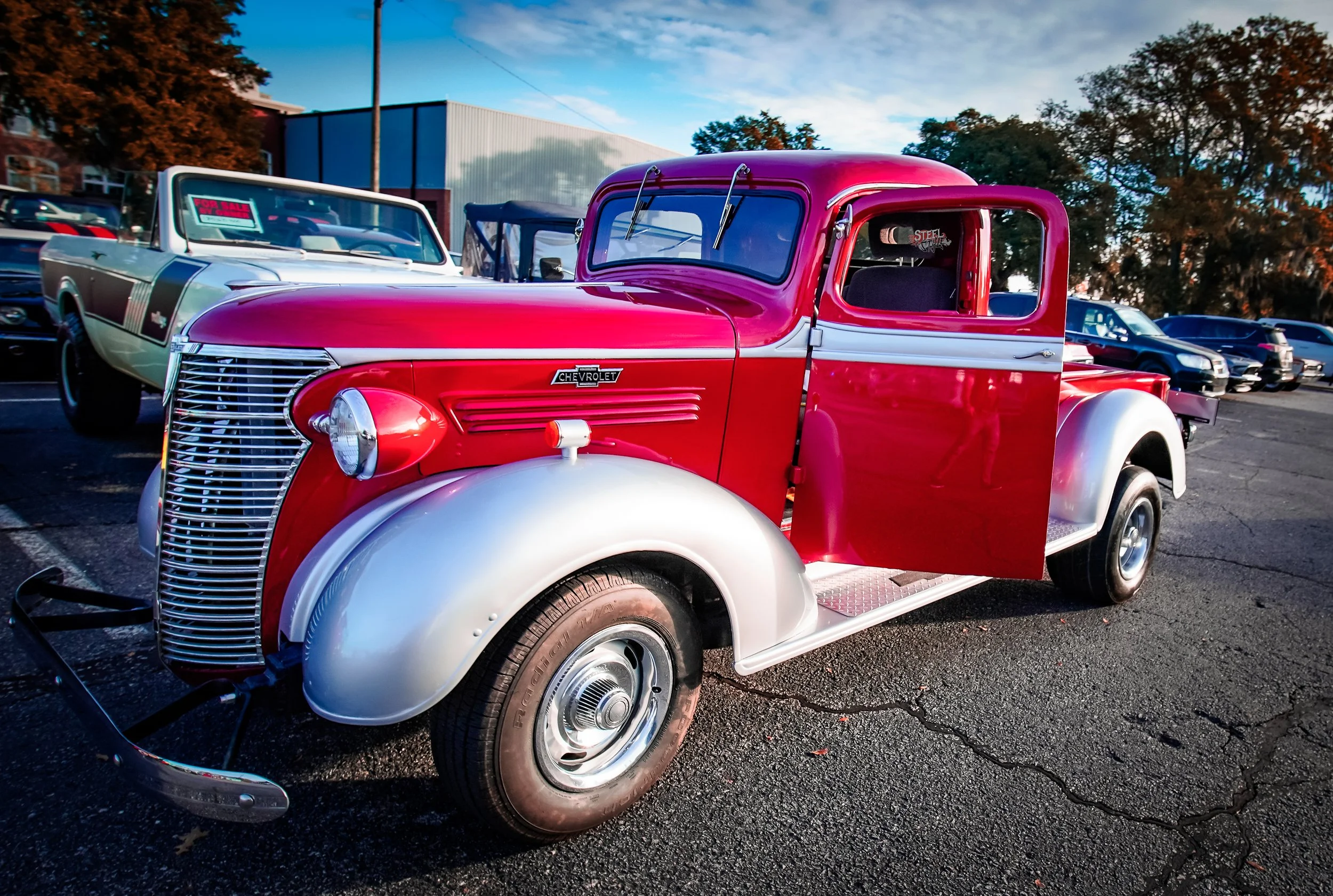 A vintage red and white Chevrolet pickup truck parked in a lot, with a row of other cars and trees in the background.
