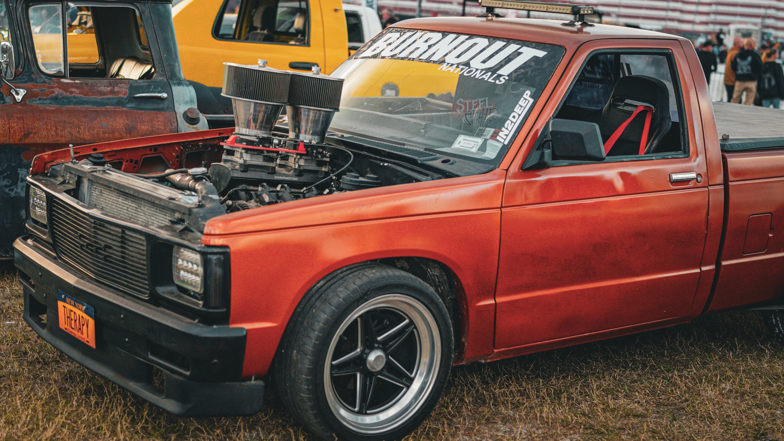 Red vintage pickup truck with modified engine and large air intake, displayed at a car show with other vehicles and people in the background.