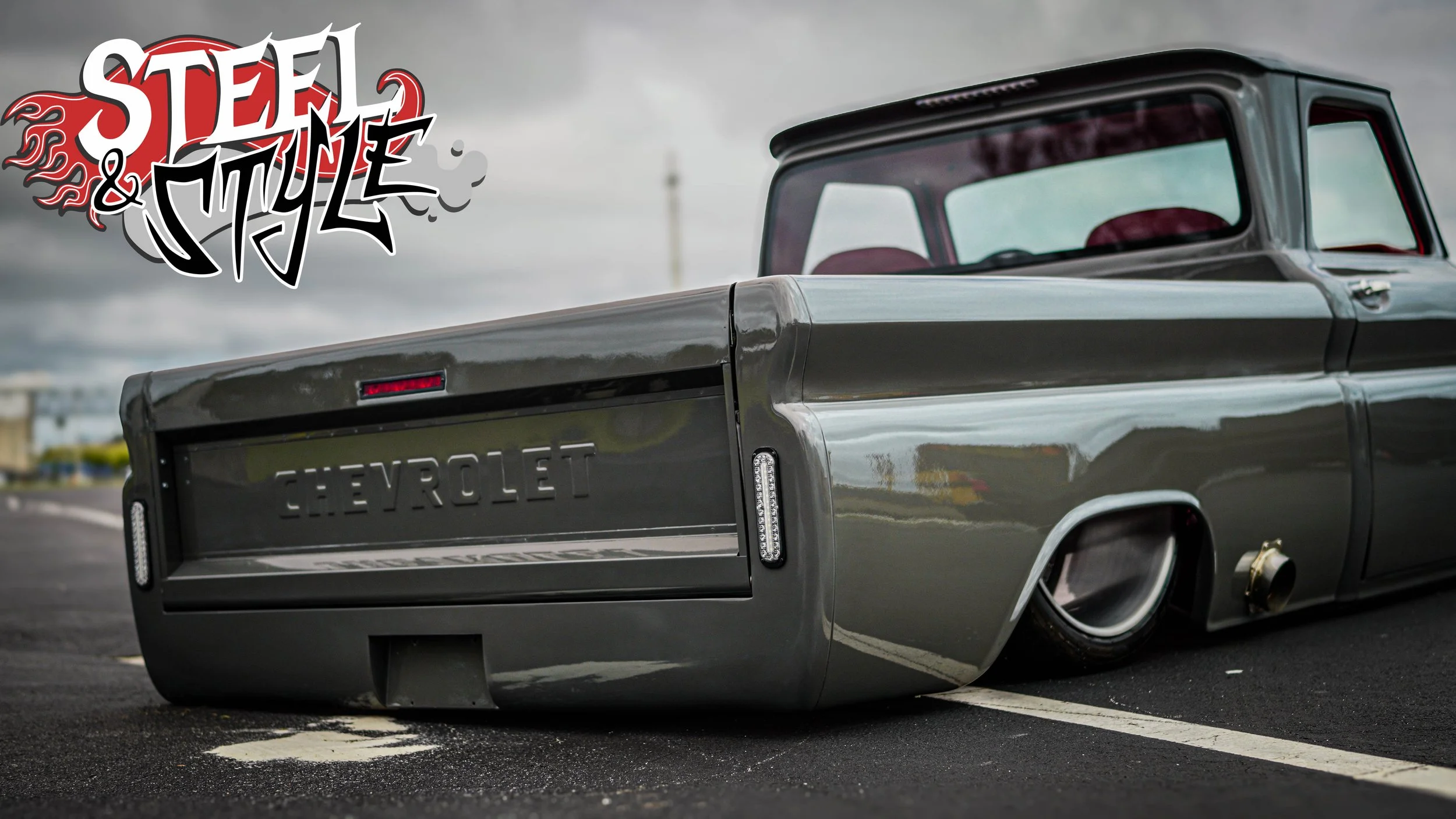 Low-riding black Chevrolet pickup truck with a custom chrome grille and red interior, parked on a dark asphalt lot on a cloudy day, with a front-oriented graffiti-style logo reading 'Stee & Miley' in the top left corner.