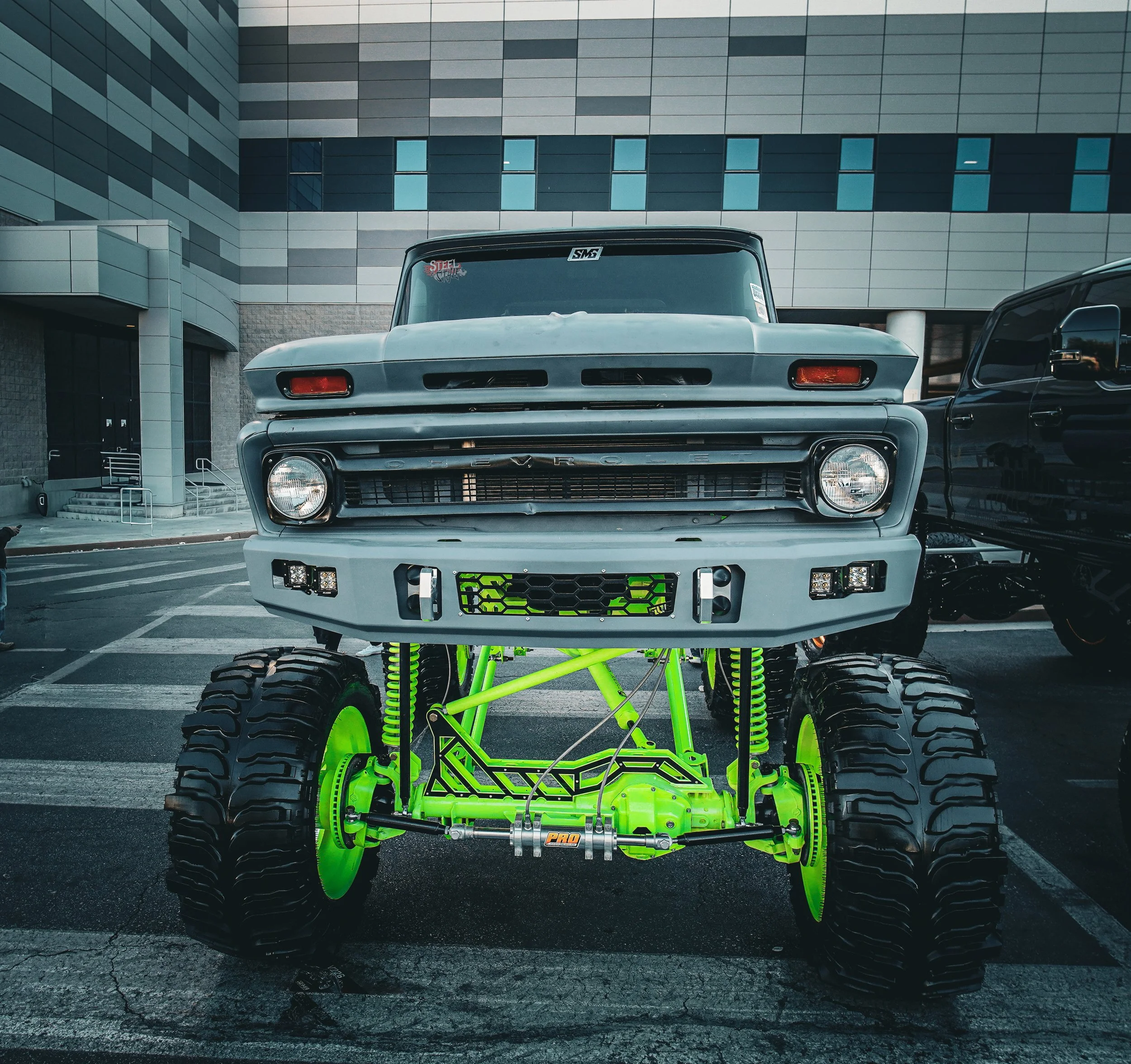 A lifted off-road vehicle with large tires, bright green suspension, and a modified front grille parked in an urban environment.