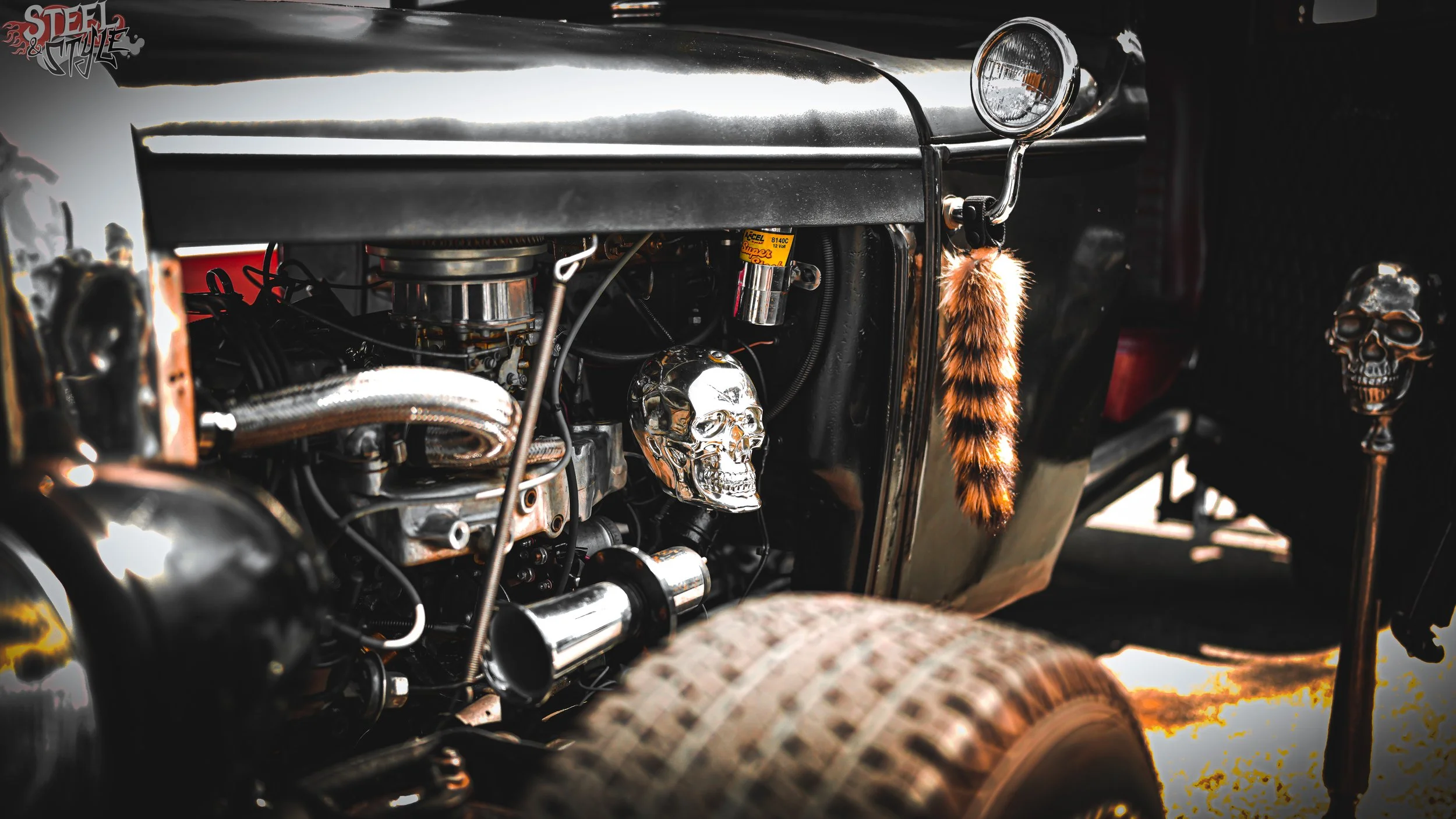Close-up of a vintage car engine decorated with chrome skull ornaments, with a tiger fur strip hanging beside the headlight.
