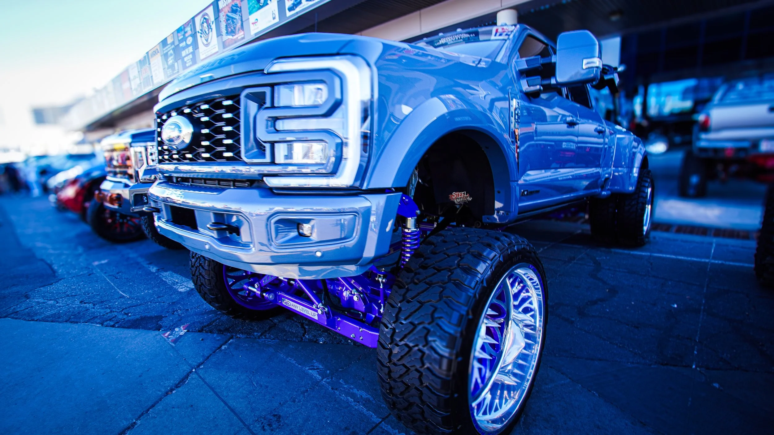 A modified blue Ford pickup truck with large tires and lifted suspension, displayed at an outdoor auto show.