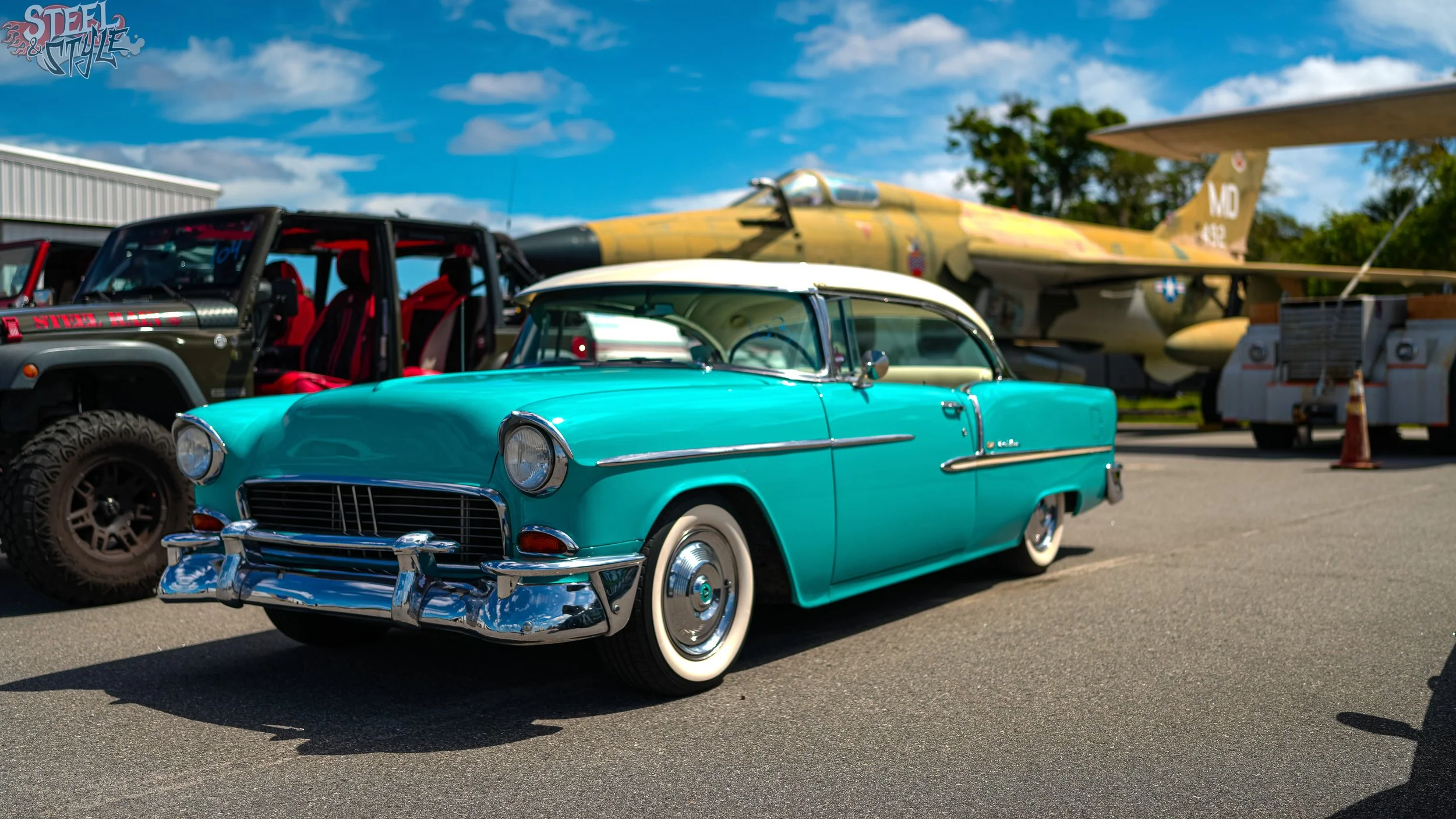 A vintage turquoise and white Thunderbird car parked in front of a military jet plane at an outdoor event.