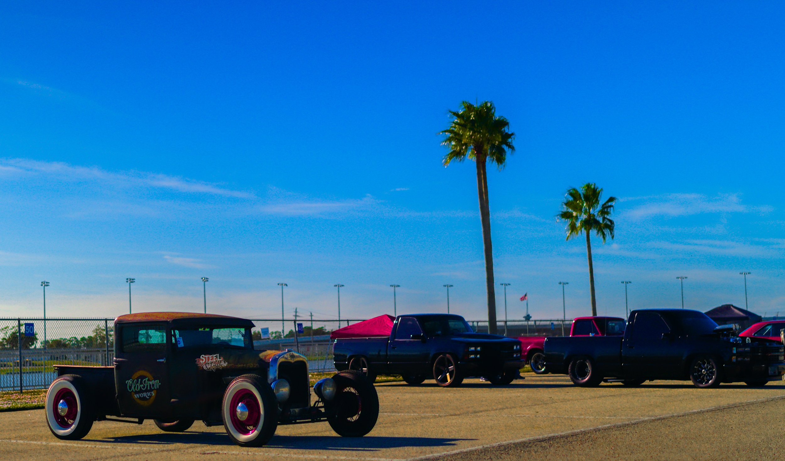 Vintage black hot rod car with pink rims parked on a dirt surface at a car show, with palm trees and other classic cars in the background under a clear blue sky.