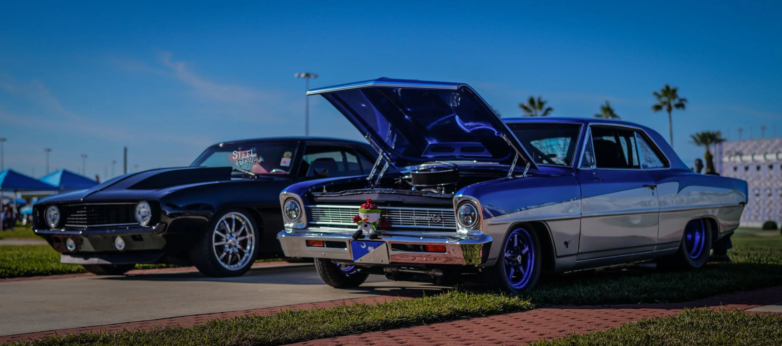 Two vintage cars parked outdoors under a clear blue sky, with one car's hood open displaying the engine, and a small stuffed toy attached to the front of the selected car. Tents and palm trees are visible in the background.