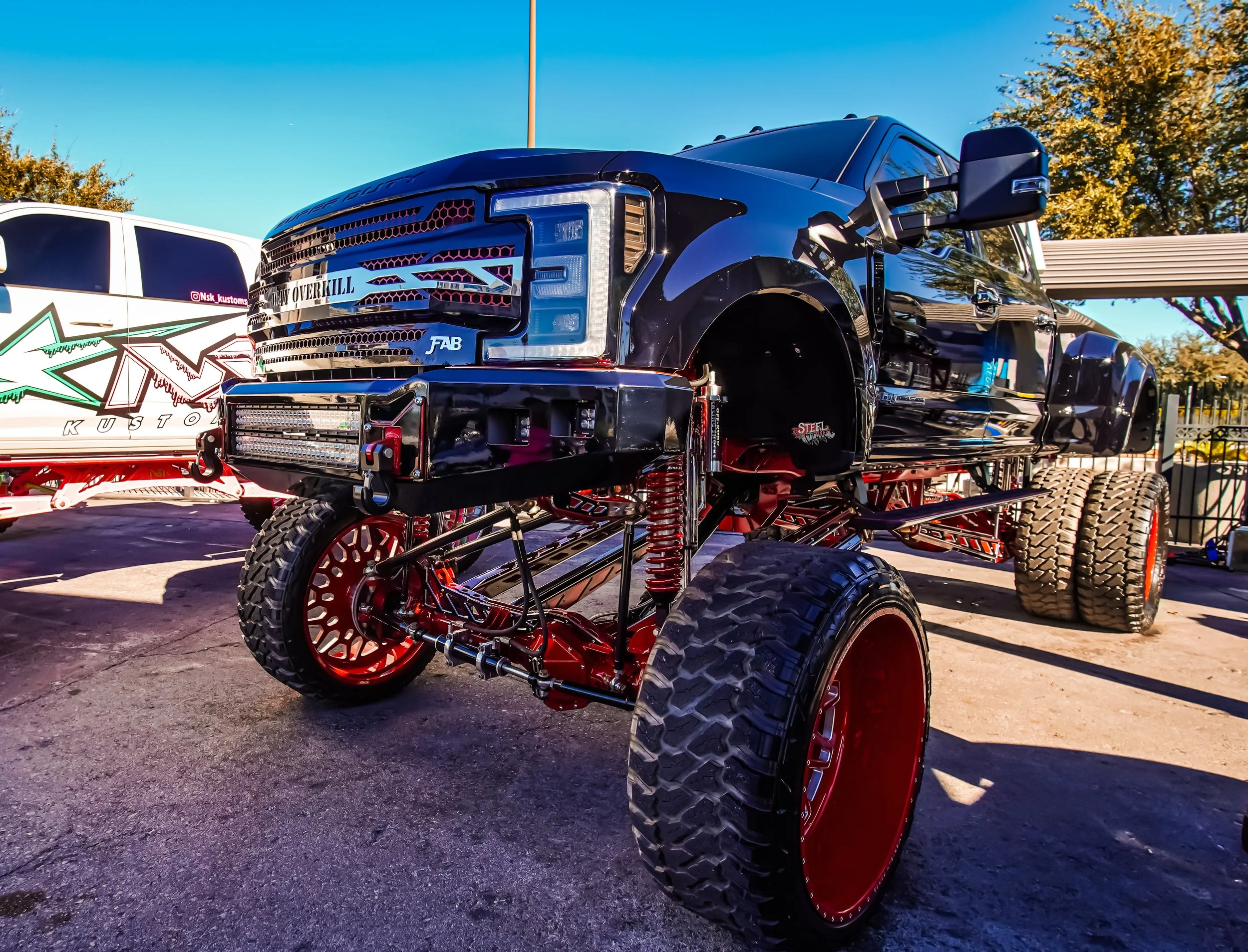 A black monster truck with large tires and red suspension components, parked on a concrete surface under a clear blue sky.
