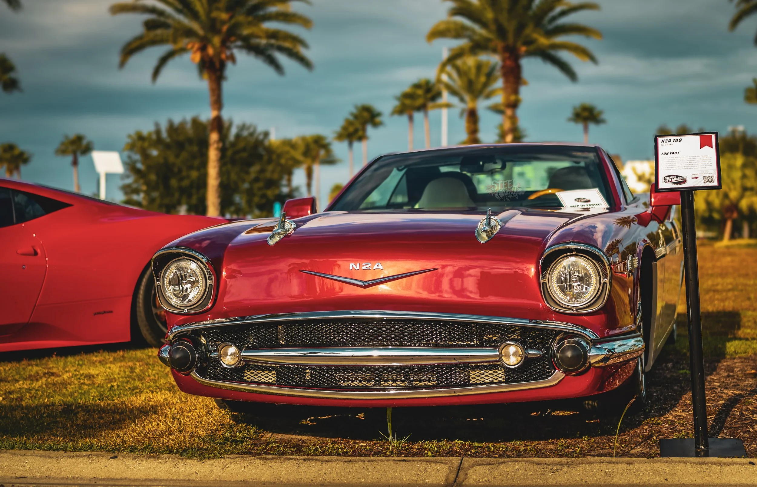 A vintage red sports car displayed at an outdoor car show, with a signboard beside it providing information.
